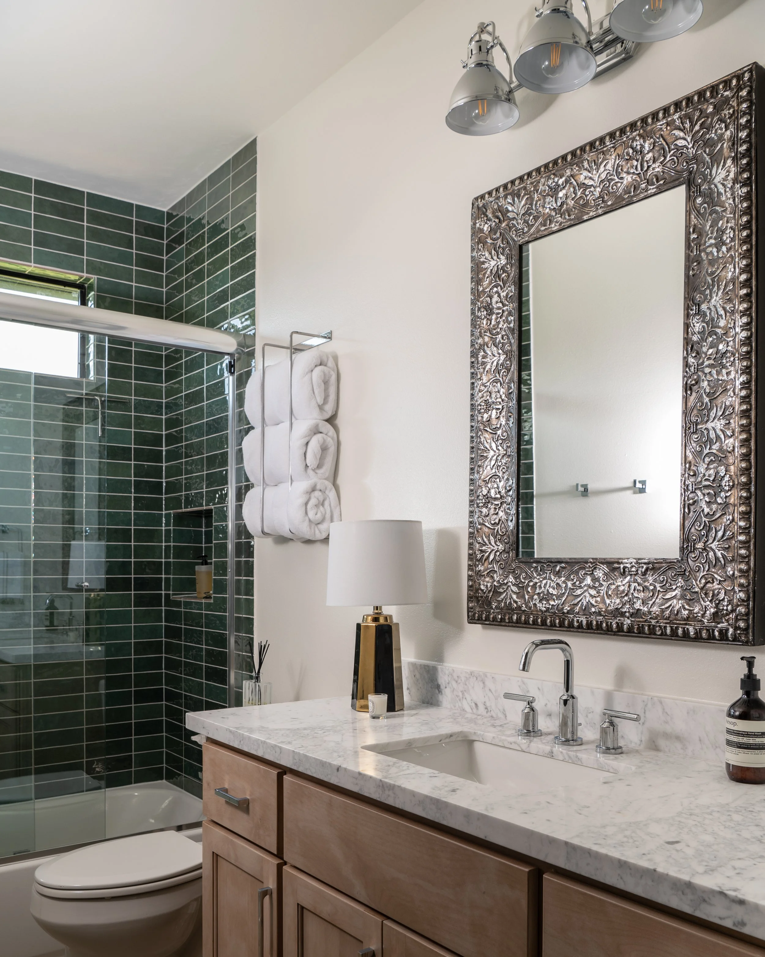 A modern bathroom featuring a wooden vanity with a marble countertop, a white sink, a decorative framed mirror, a silver light fixture with three lamps, a table lamp, a towel rack with rolled white towels, and a shower area with dark green tiles and 
