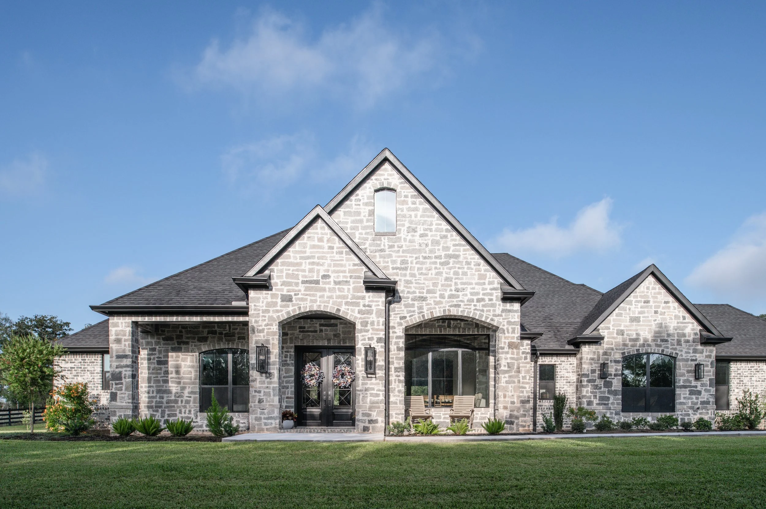 A large house with stone exterior and black roof, featuring a front porch with two chairs and decorated doors, under a blue sky with some clouds.