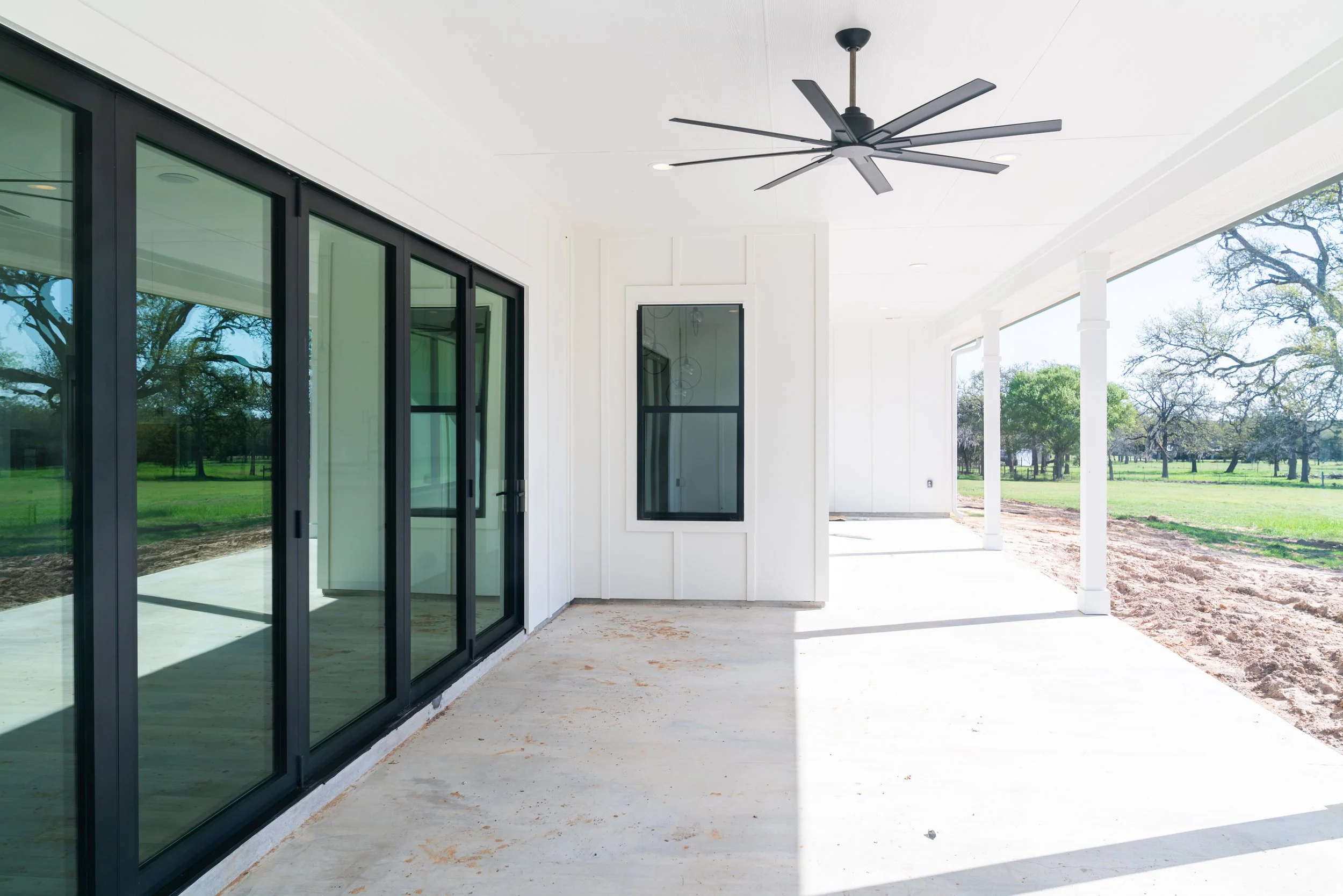 Empty covered patio with black-framed sliding glass doors, white walls, and a ceiling fan, overlooking a grassy yard with trees.