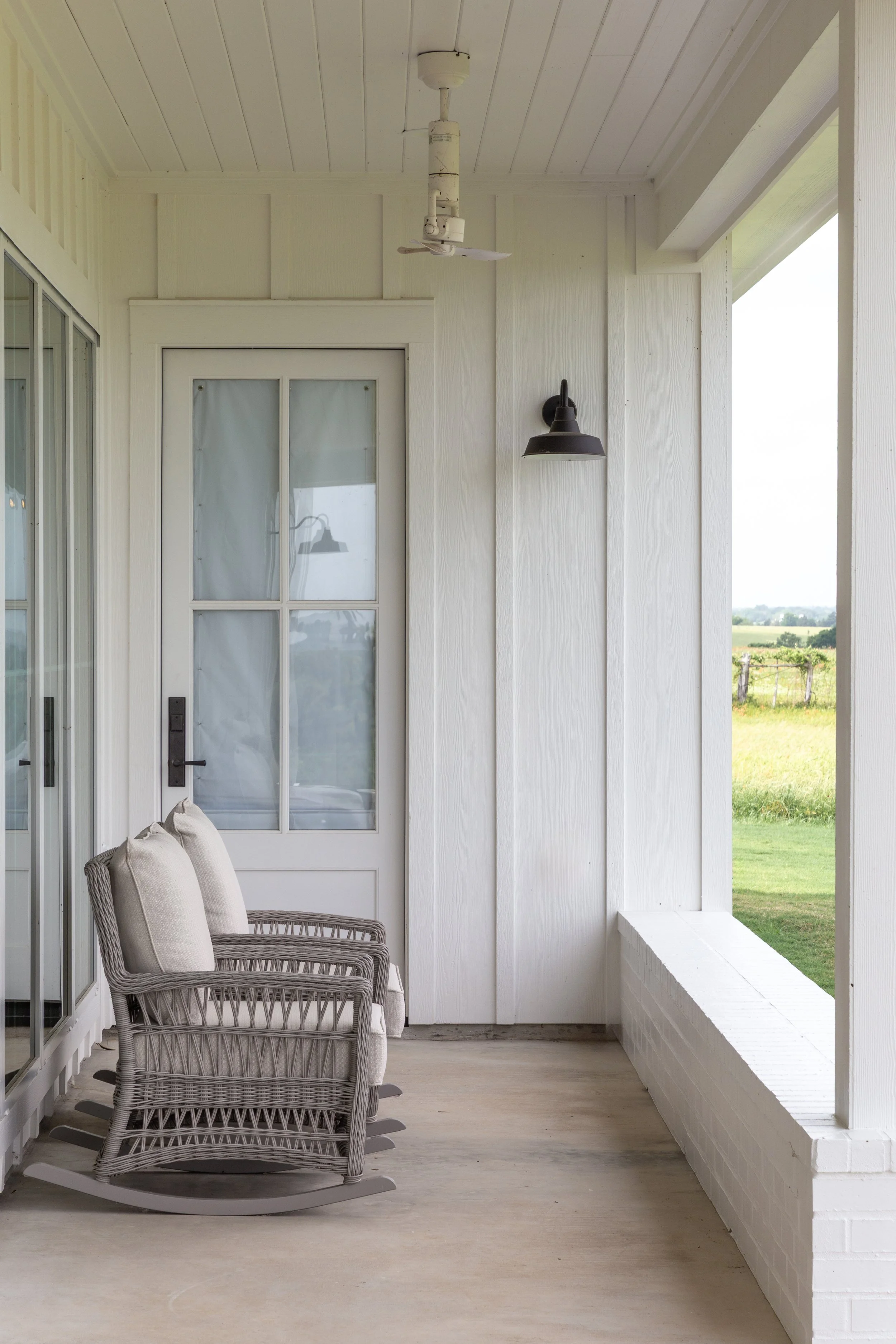 Porch with two wicker rocking chairs with cushions, white station door with glass panes, black wall sconce, ceiling fan, and grassy field view in the background.