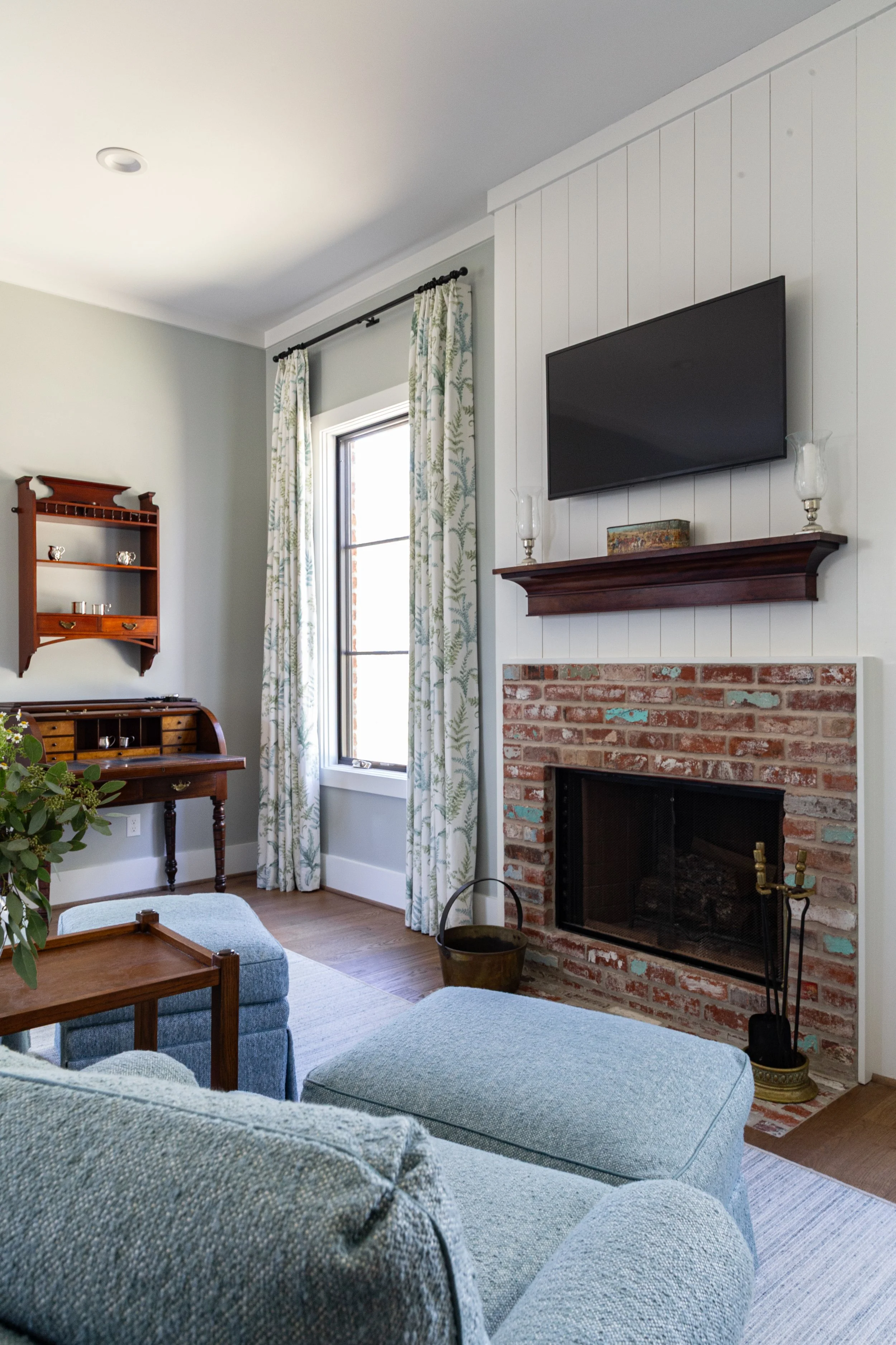 Living room with a brick fireplace, a mounted flat-screen TV, a window with patterned curtains, a blue sofa, and a small wooden writing desk with shelves.