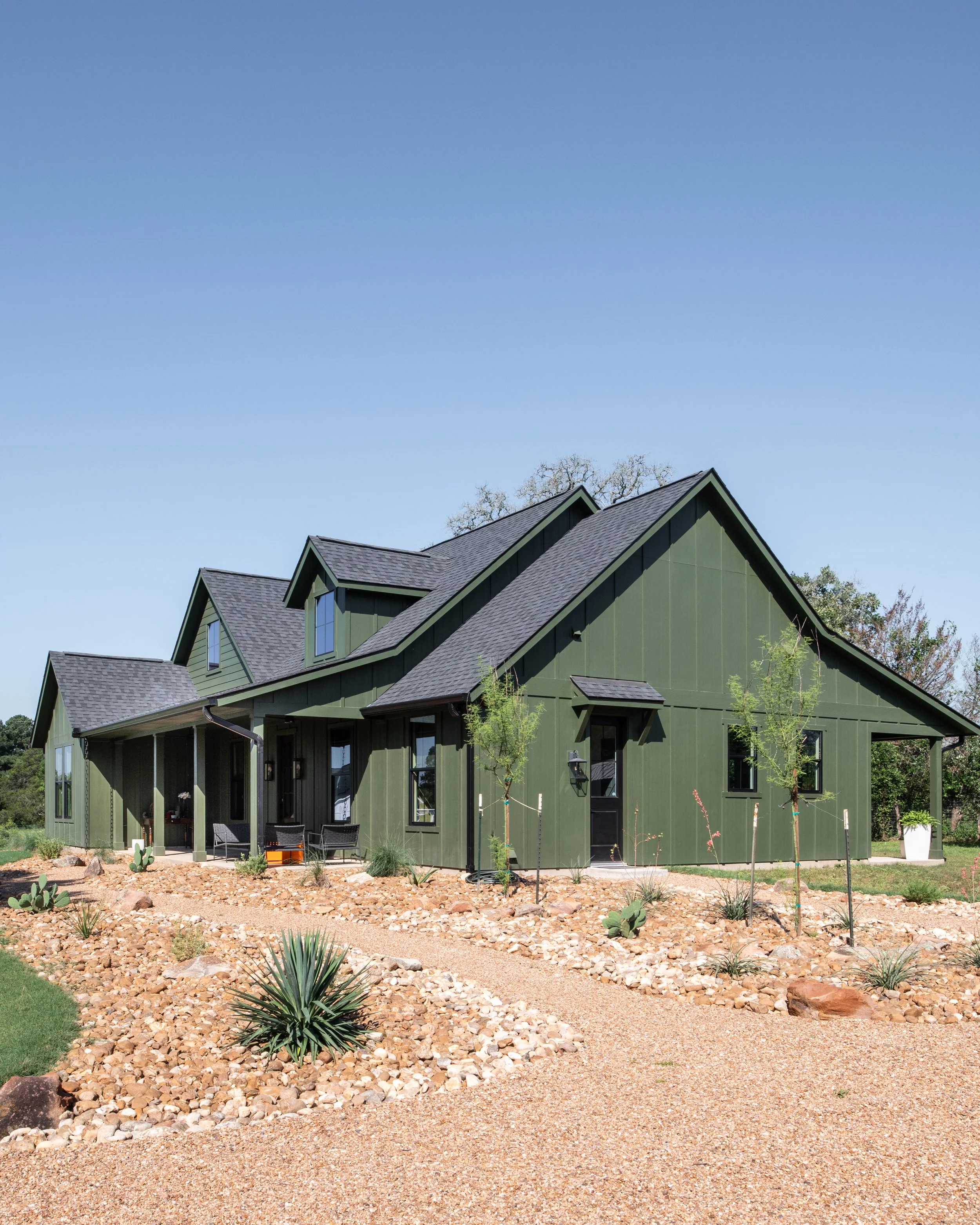 A modern green house with multiple roof peaks and large windows, surrounded by desert landscaping with rocks, small trees, and cacti.