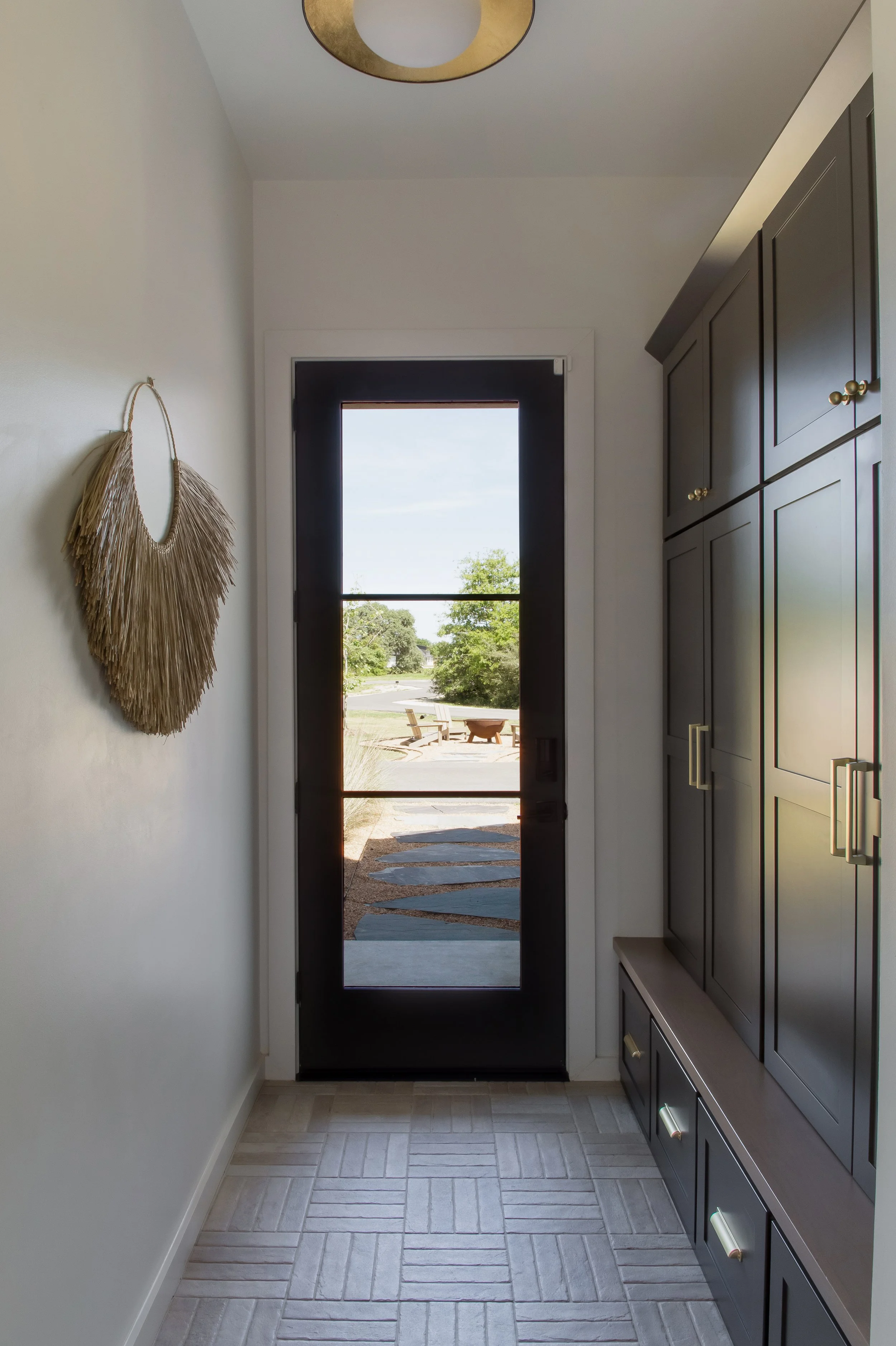 A modern entryway with a black front door and a glass window, view of a patio with a fire pit and outdoor seating, dark gray cabinets on the right, and a decorative wall hanging on the left.