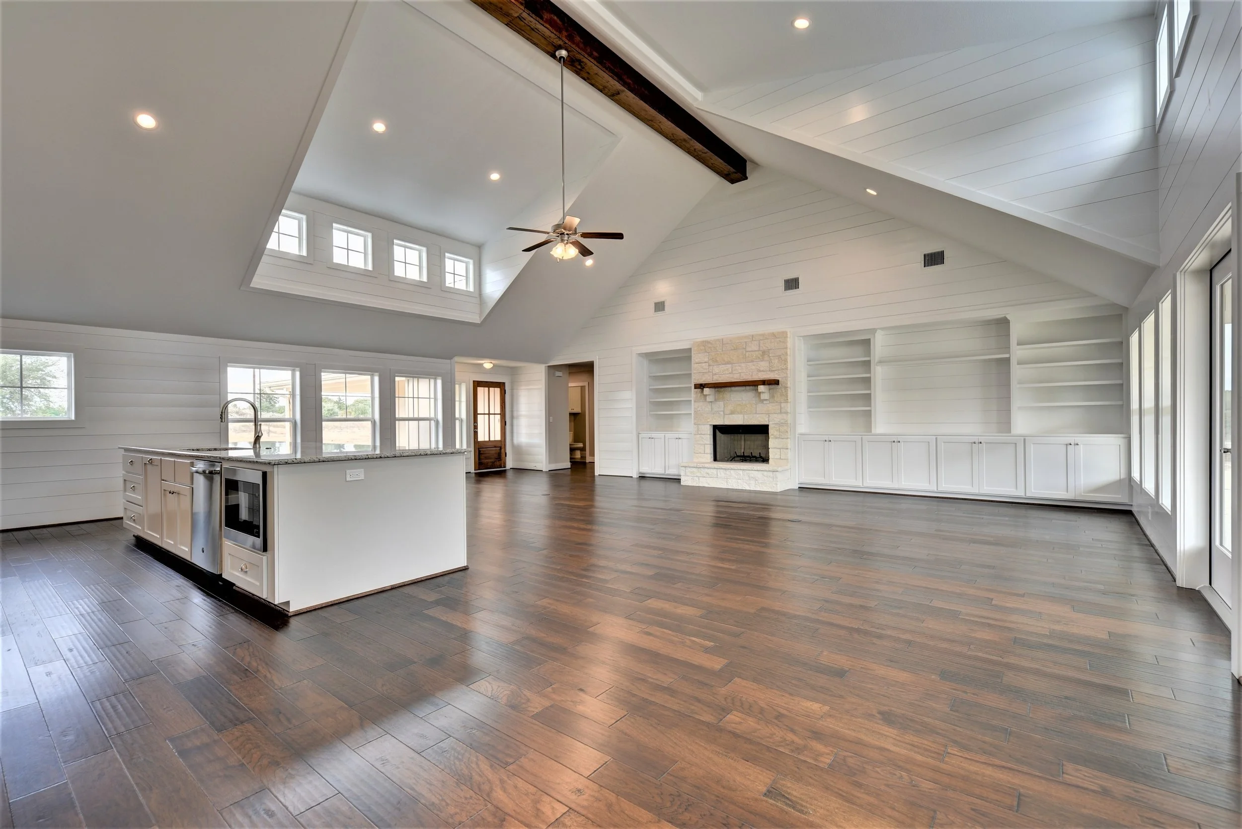 Empty living room with white walls, wood floor, fireplace, built-in shelves, and kitchen island. Large windows and skylight bring natural light.