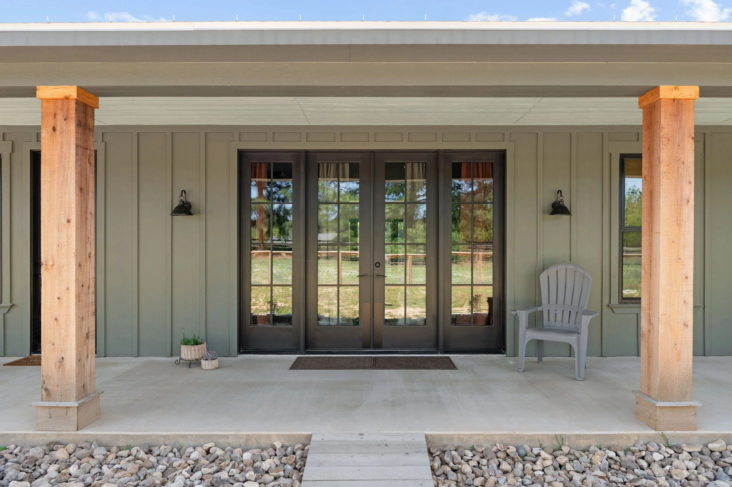 Front porch with double glass door, two black wall-mounted lamps, a white plastic armchair, small pot with plants, and pebble landscaping.