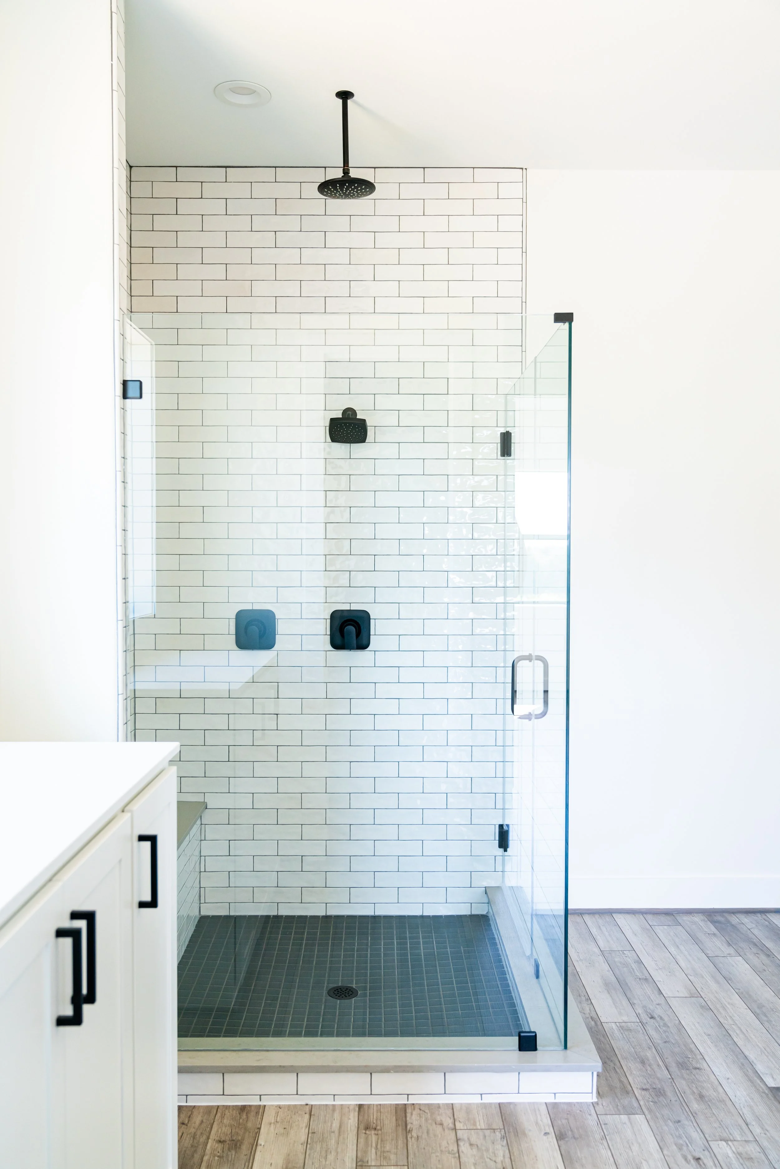 White-tiled walk-in shower with black fixtures and a glass door, in a modern bathroom with wood flooring.