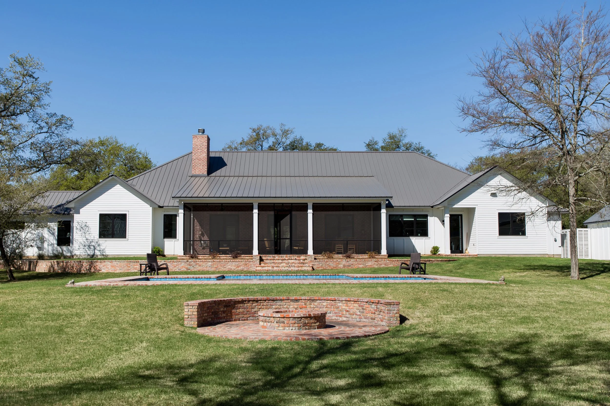 Backyard view of a large white house with a gray metal roof, surrounded by green grass, trees, and a blue sky. There is a brick firepit in the foreground and a swimming pool with lounge chairs. The house has multiple windows and a screened porch.