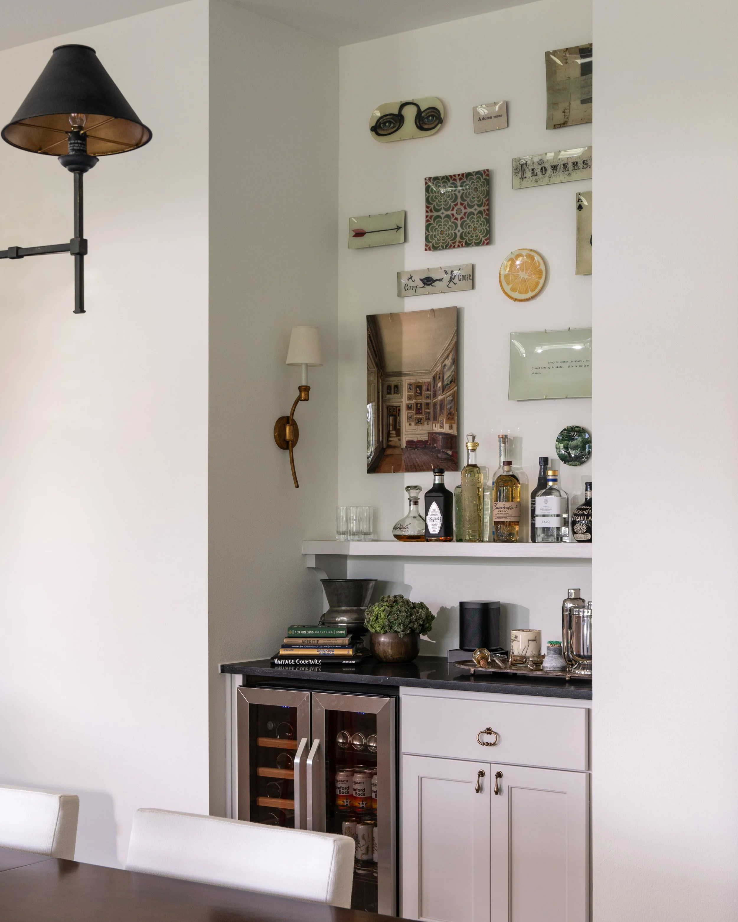A room with a white wall decorated with various pictures, signs, and a mirror. A white cabinet with a black countertop holds liquor bottles, glasses, a plant, and books. Part of a dining table with white chairs is visible in the foreground. A black w