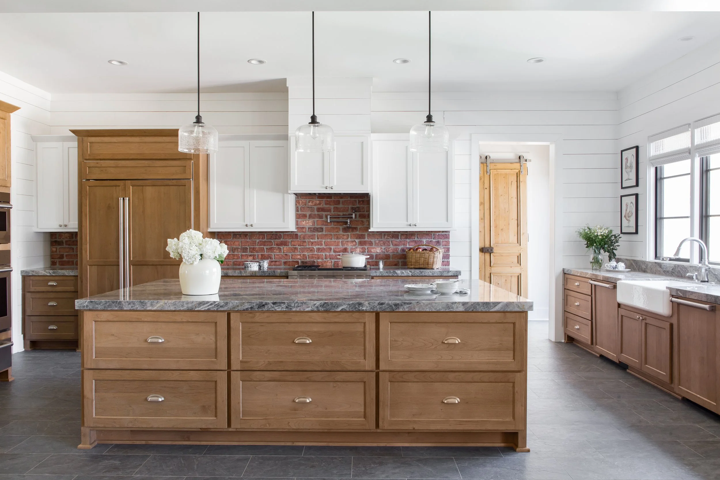 Modern kitchen with white upper cabinets, wooden lower cabinets, a large wooden island with a marble countertop, a brick backsplash, three pendant lights above the island, a vase with flowers, and a farmhouse sink near a window.