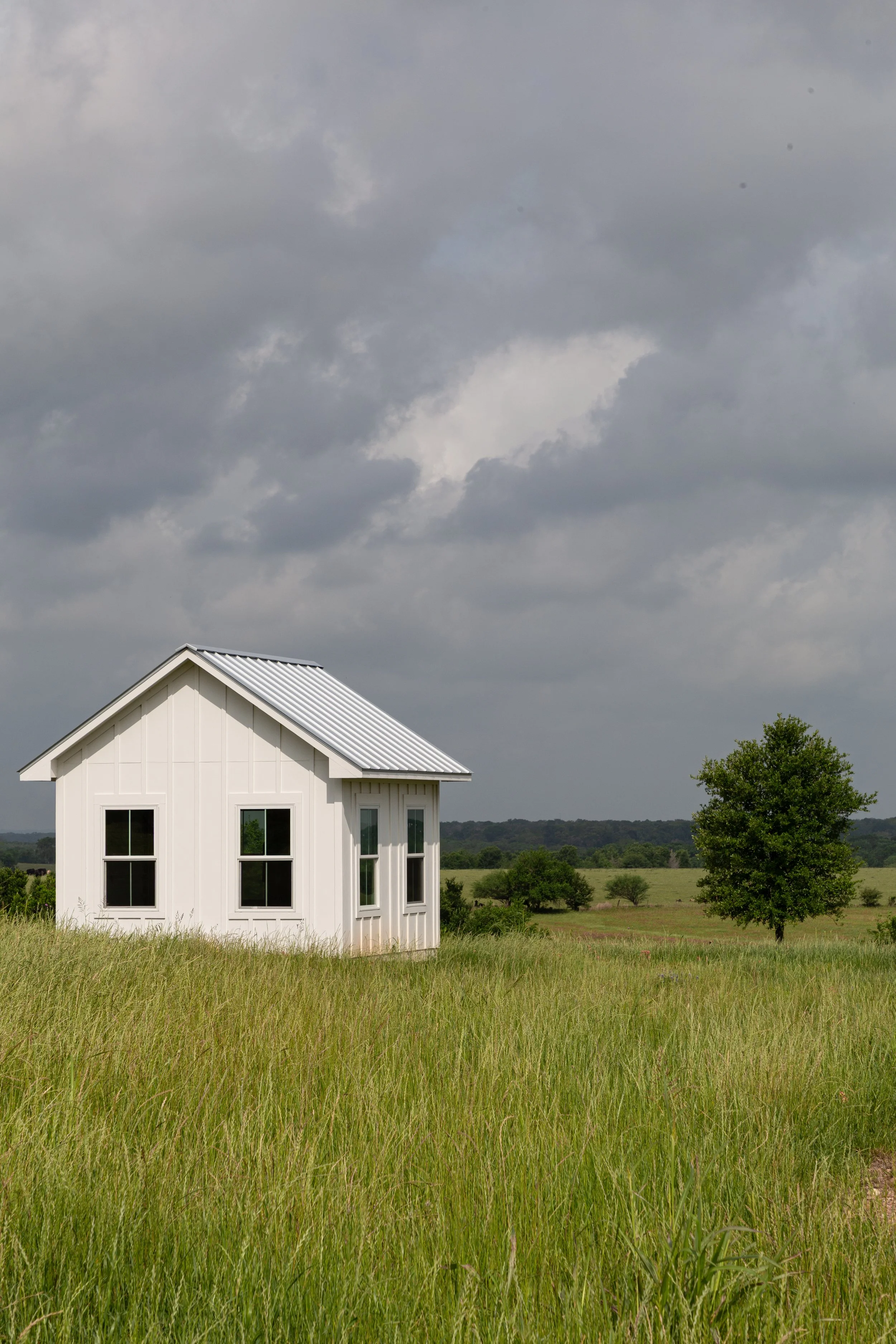 A small white house with a metal roof sits in a green field with tall grass and a few trees in the background under a cloudy sky.