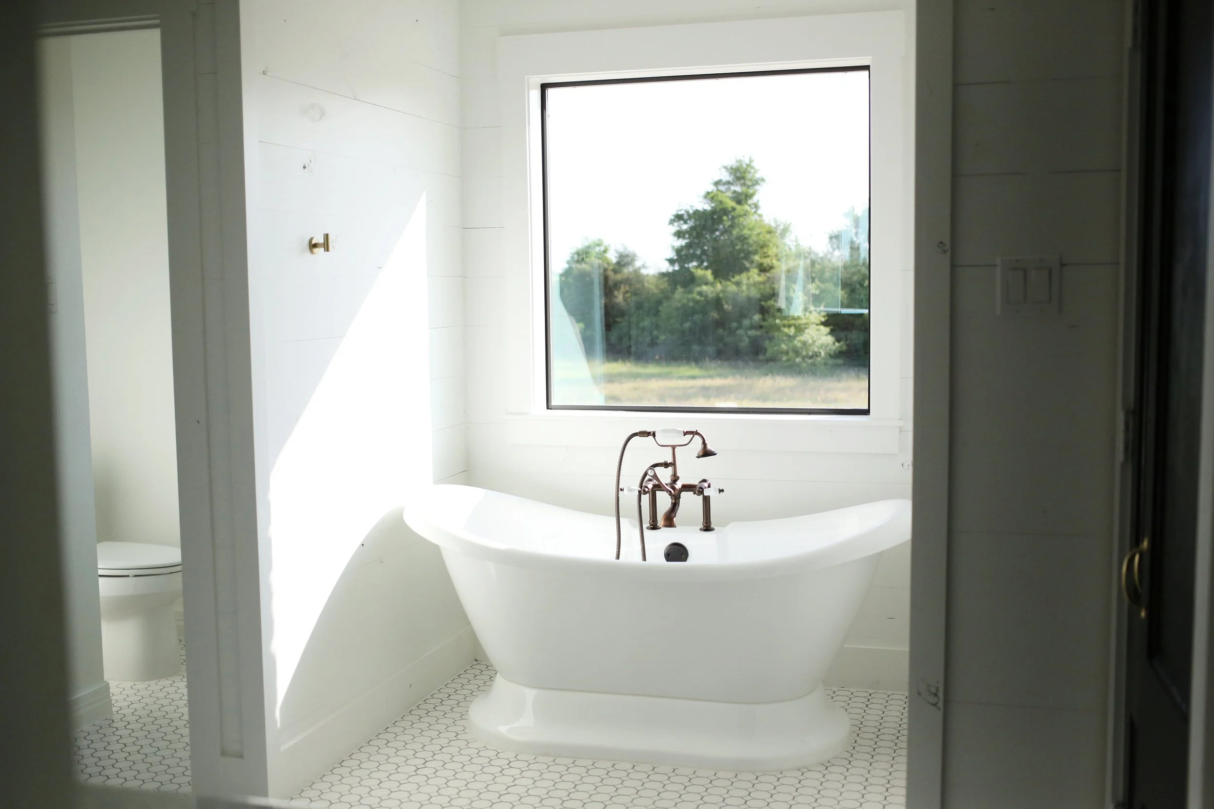 White bathtub with bronze faucet beside a large window showing greenery outside in a bathroom.