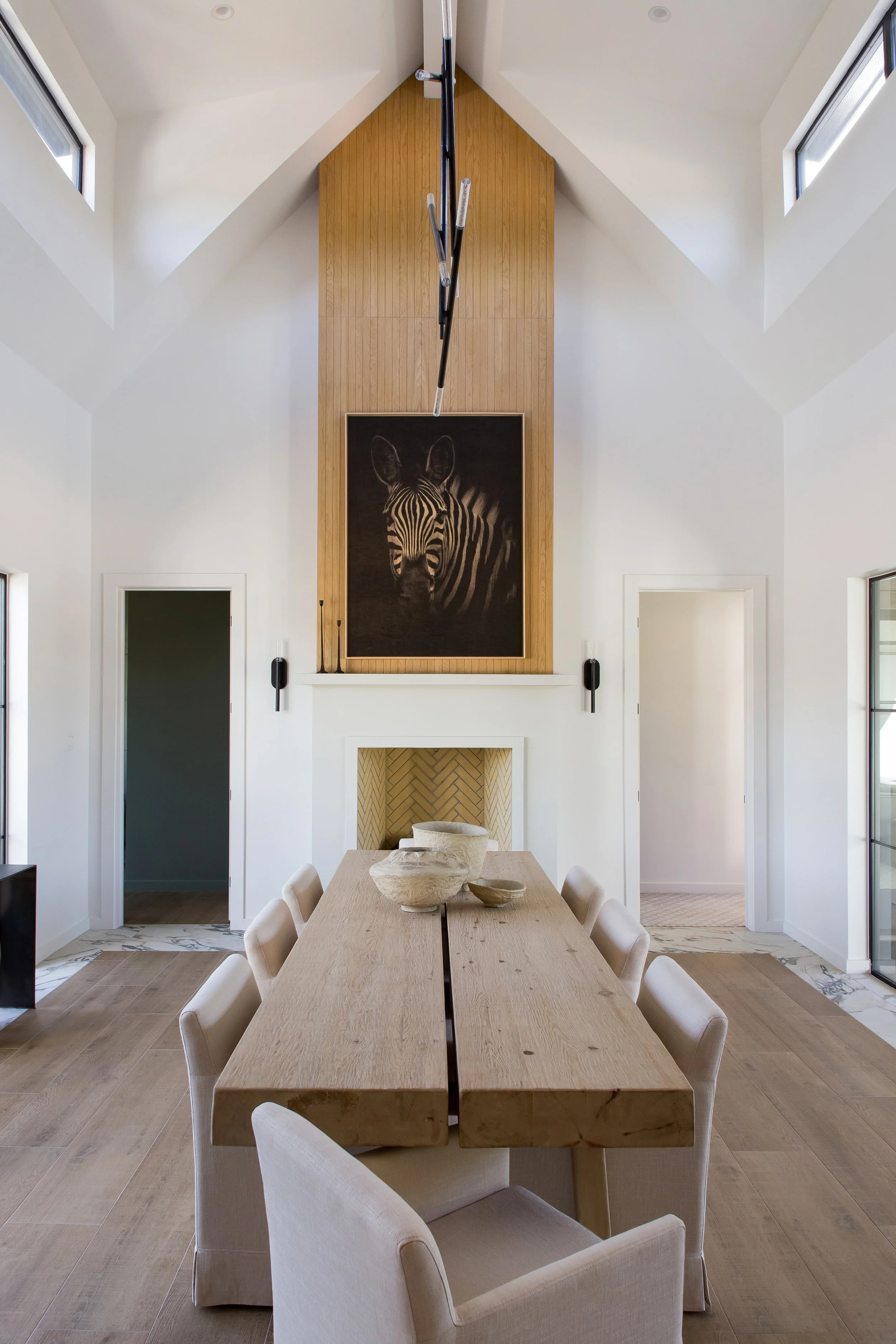 Modern dining room with a long wooden table, white chairs, large windows, light wood flooring, a fireplace with a built-in surround, and a large artwork of a zebra on a wooden panel above the fireplace.