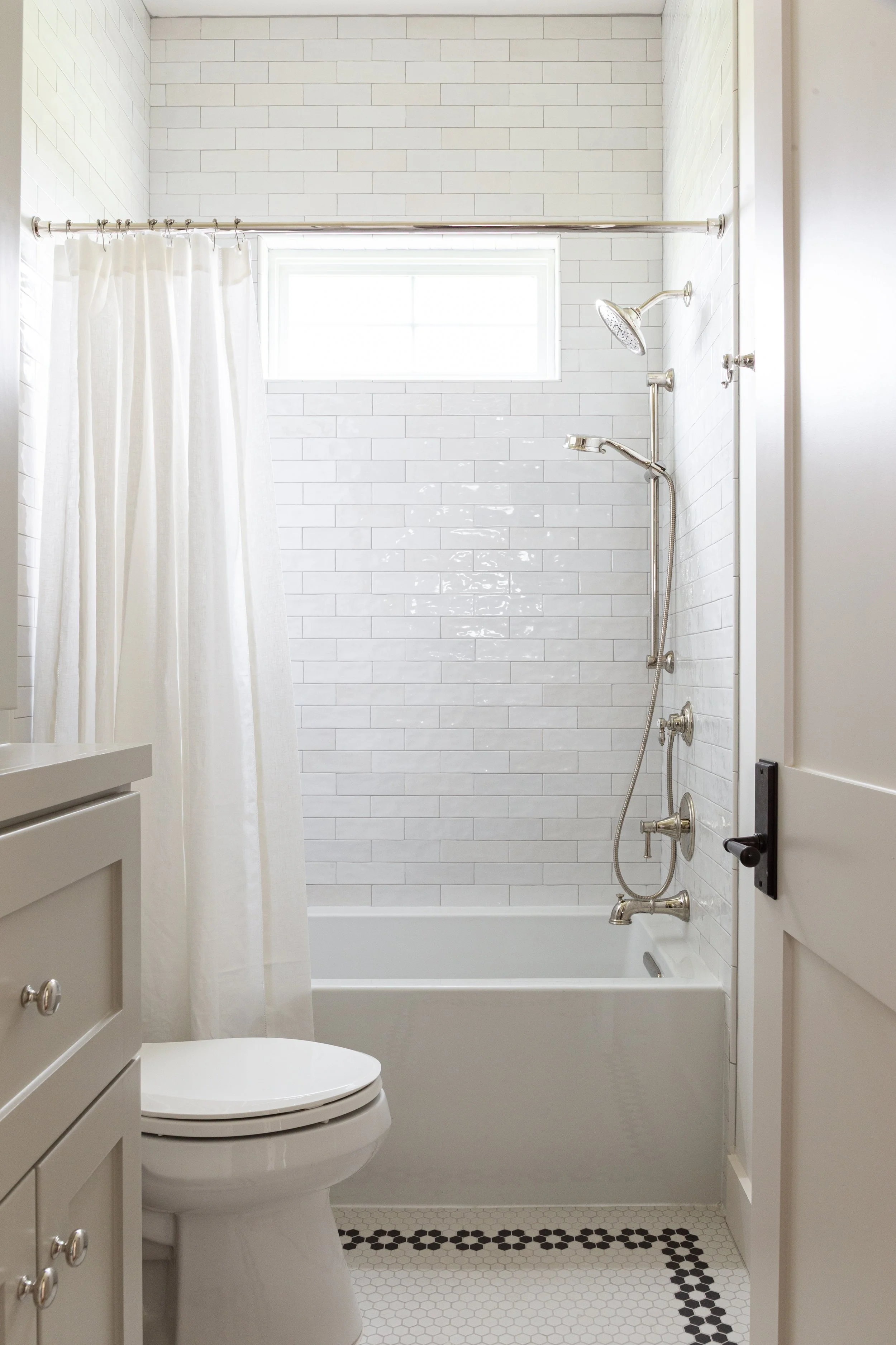 Bright bathroom with a white bathtub, showerhead, and white subway tile walls, a small window, and a curtain, with a white toilet and beige cabinetry.