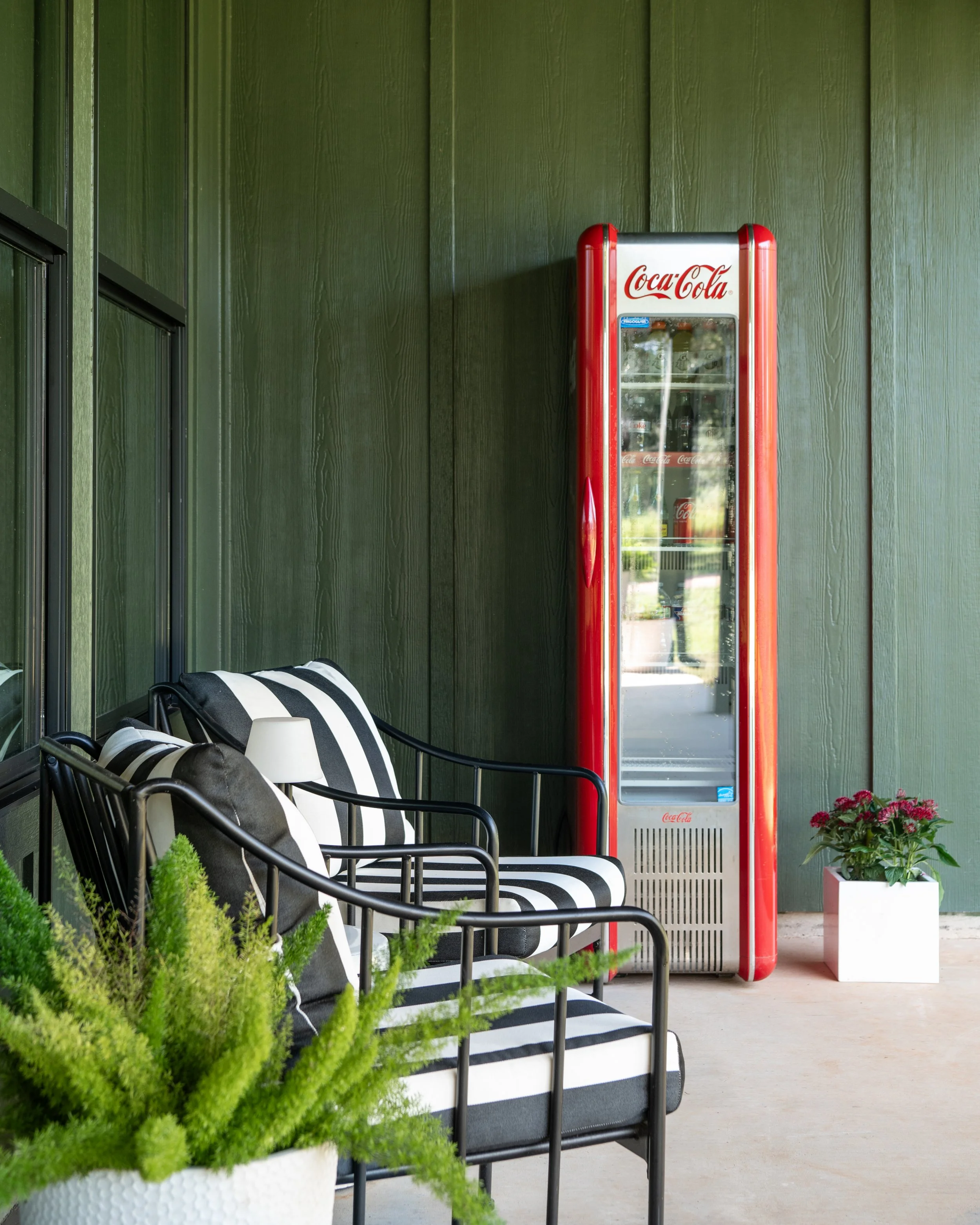 A vintage Coca-Cola vending machine standing next to a black and white striped outdoor lounge chair with pillows, a white pot with green plants, and a white pot with pink flowers, against a dark green wall.