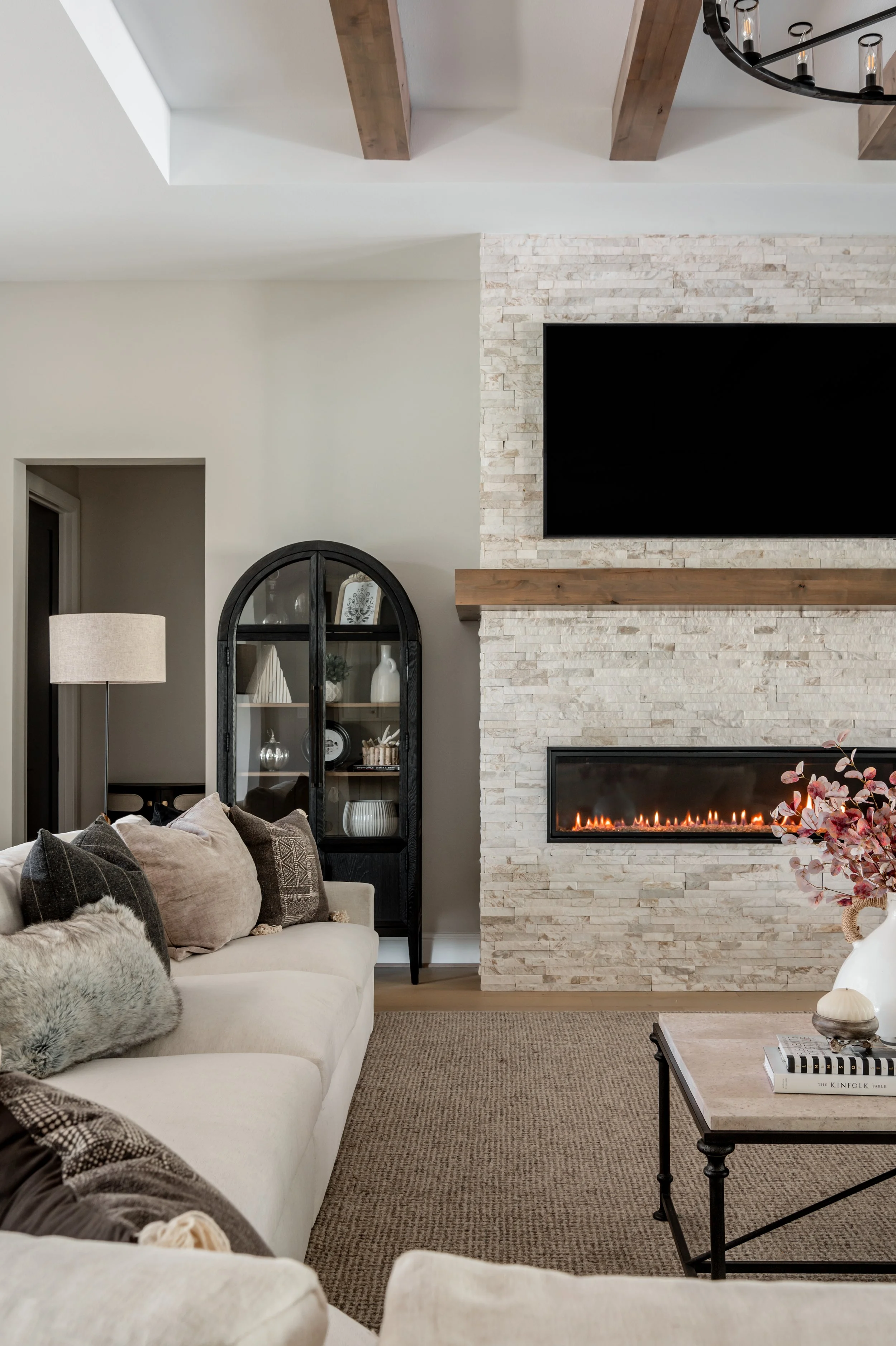Living room with a beige sofa, decorative pillows, a white planter with pink flowers, a black arched display cabinet with white and silver décor, a fireplace with a stone surround, a mounted flat-screen TV, a round black chandelier, and wooden ceilin