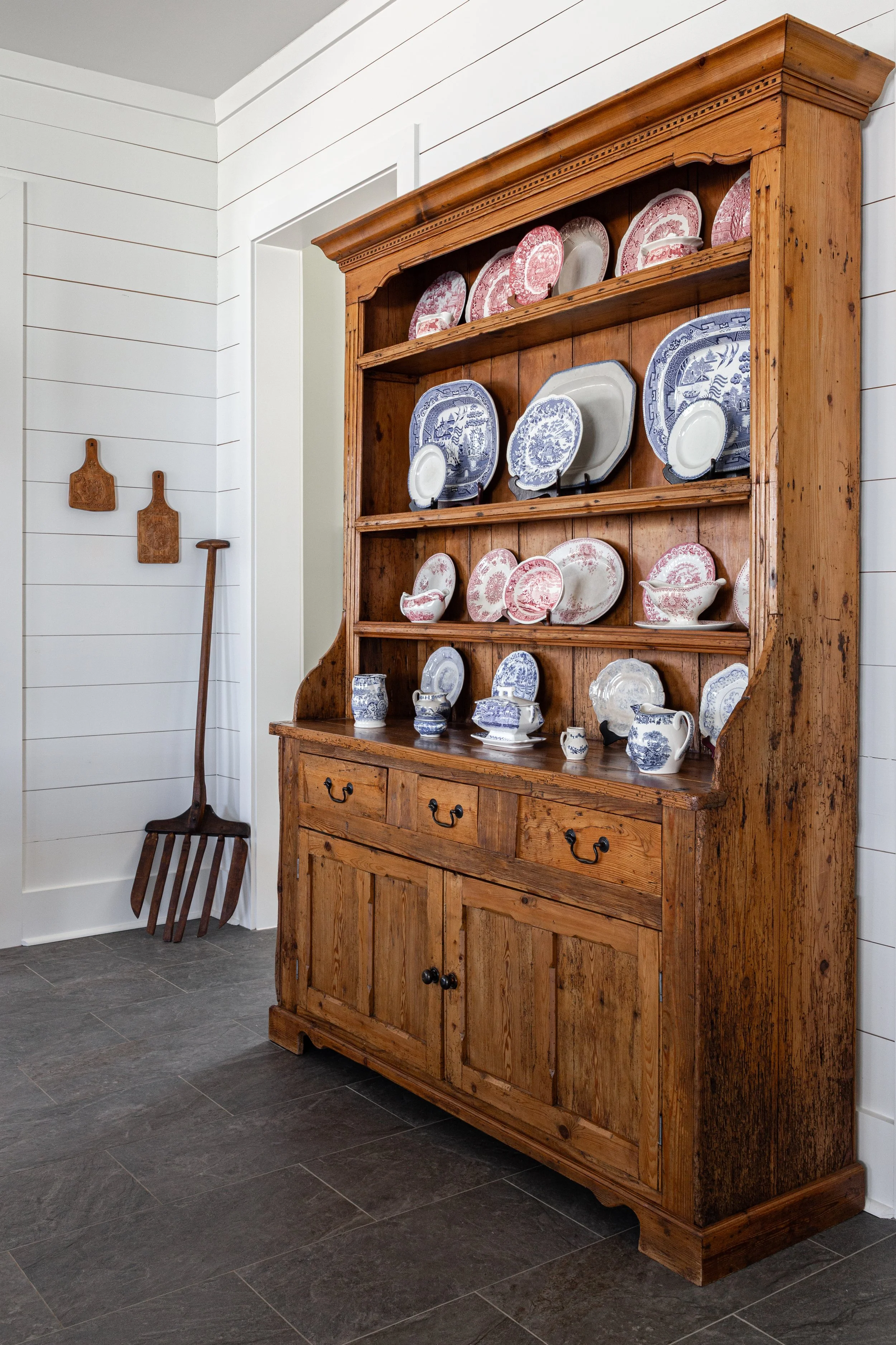 Wooden kitchen cabinet filled with decorative dishware, with a wall of white wood planks and a small wooden gardening shovel and two hooks hanging on the wall beside it.