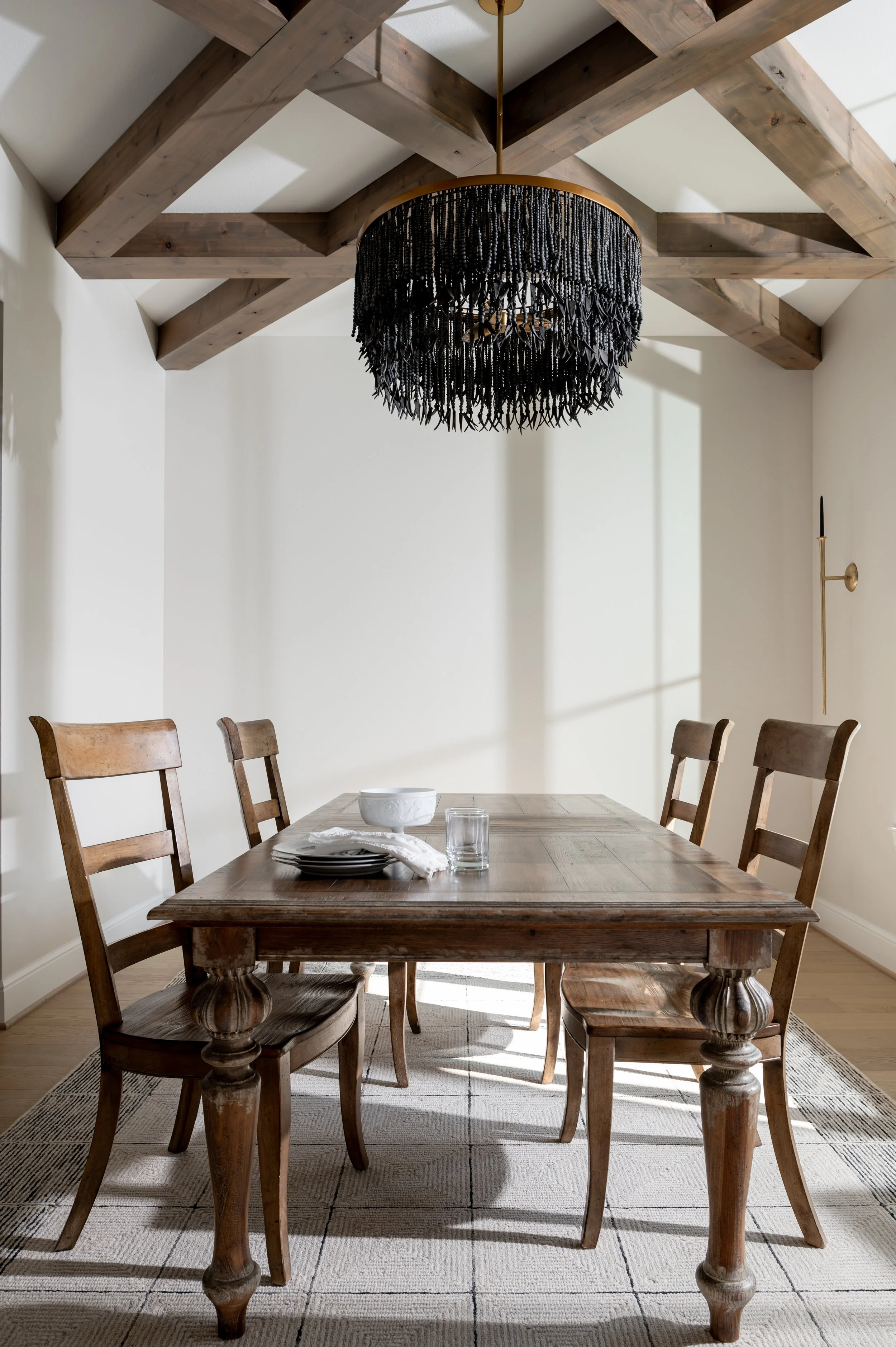 A dining room with a rustic wooden table and four matching chairs, a black chandelier hanging from a ceiling with exposed wooden beams, and sunlight casting shadows on white walls.