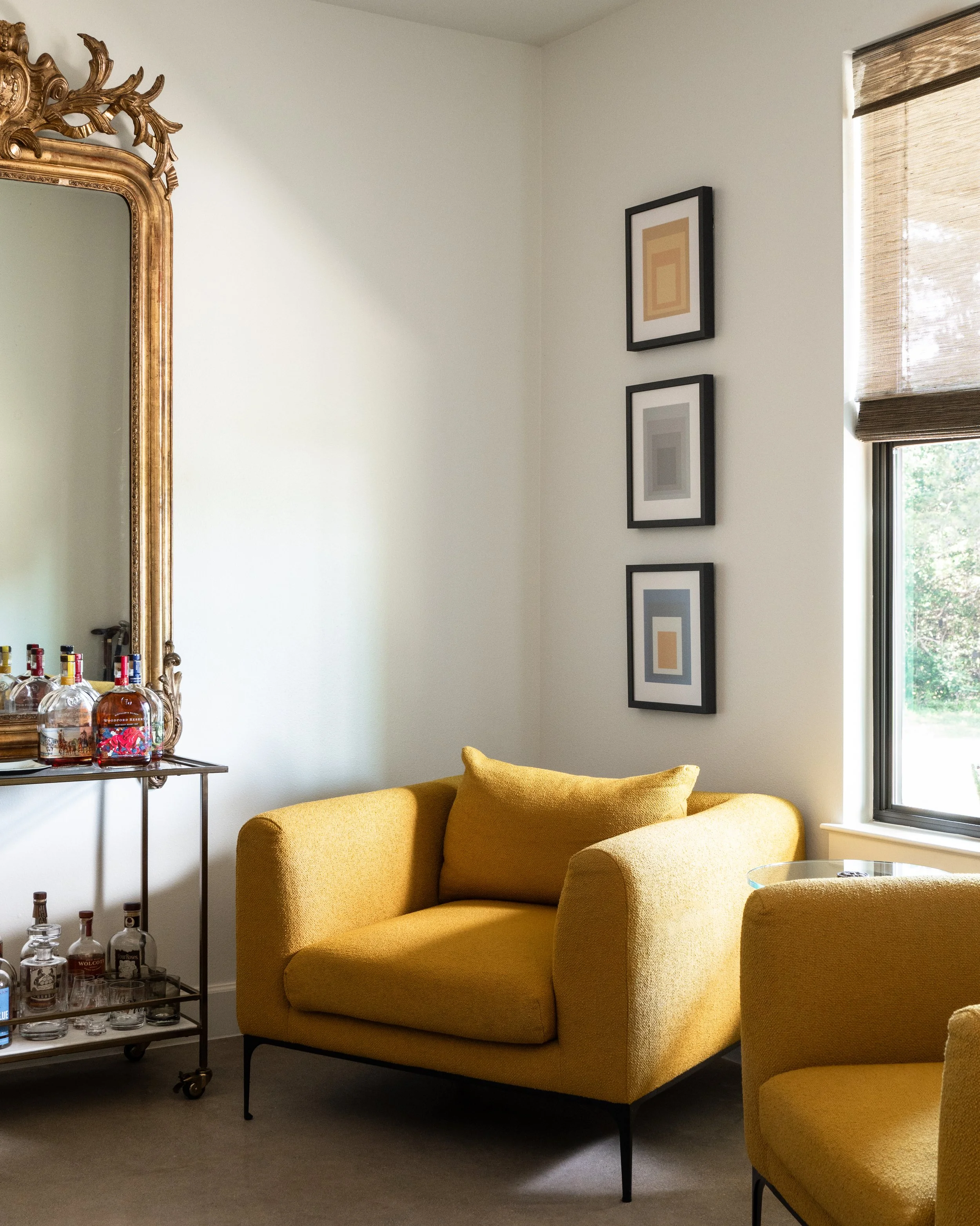 Living room with mustard yellow armchairs, a glass side table, a large ornate gold mirror, framed art on the wall, and a window with a bamboo shade.