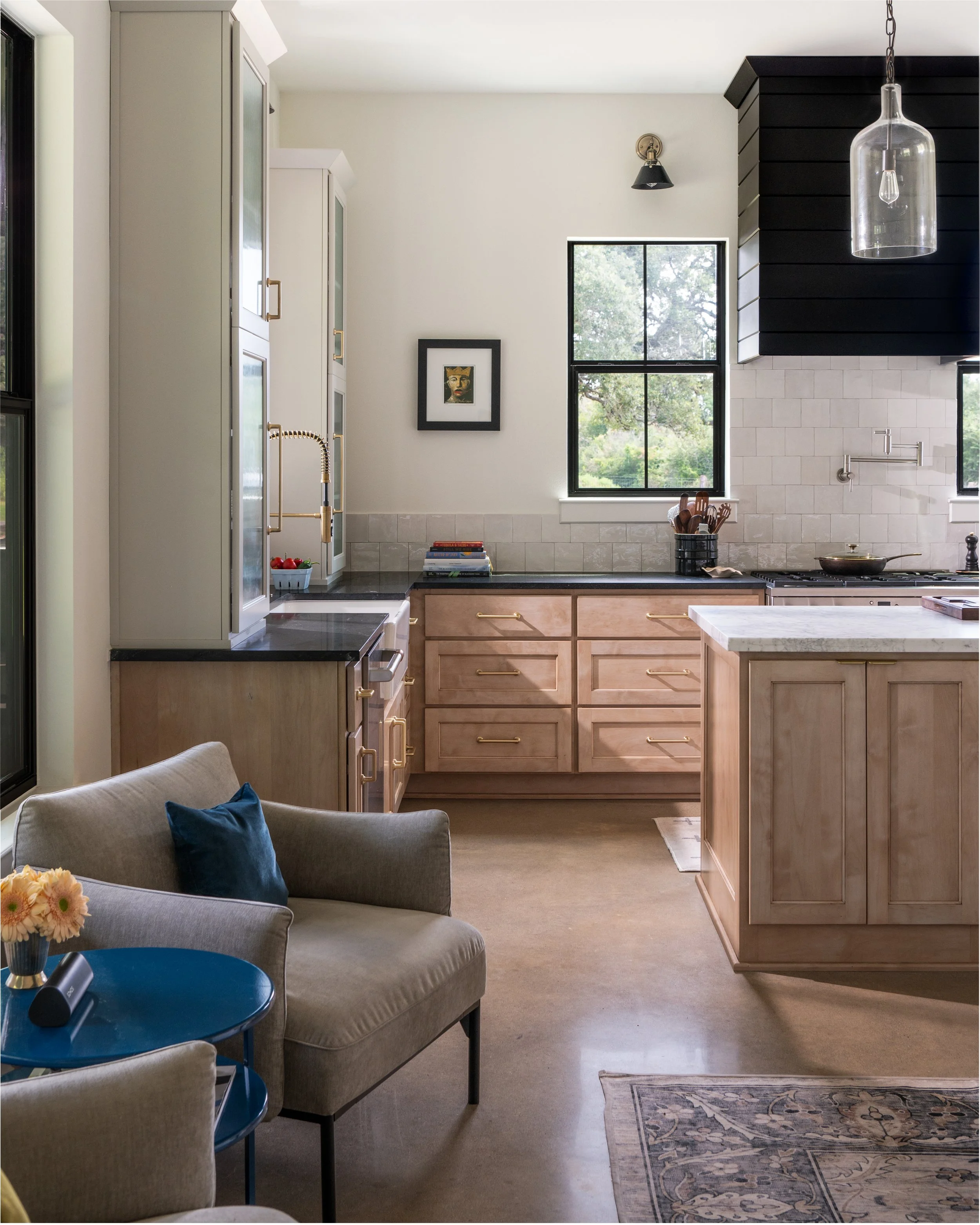 Modern kitchen with light wood cabinets, black countertops, and a black and white color scheme. Features a window looking outdoors, decorative light fixtures, and a seating area with a beige chair and blue cushion.