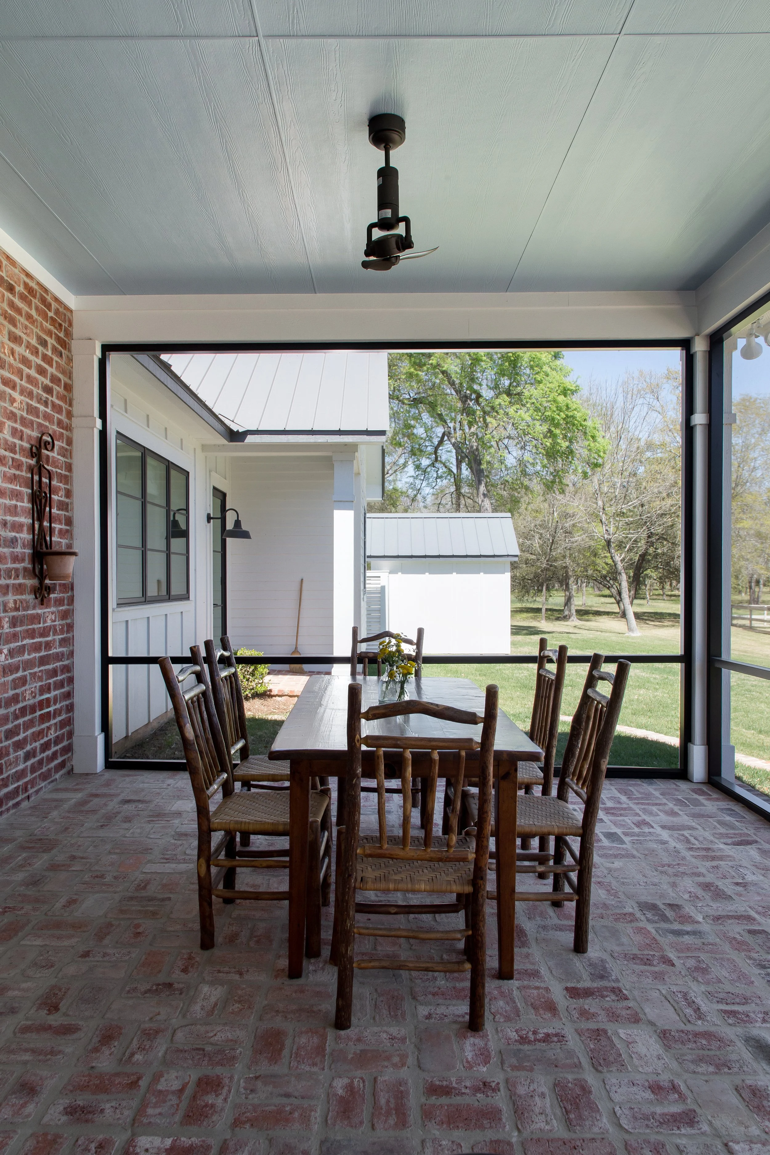 Screened porch with a wooden dining table and eight chairs, brick and white siding walls, ceiling light fixture, outside view of trees and grass.