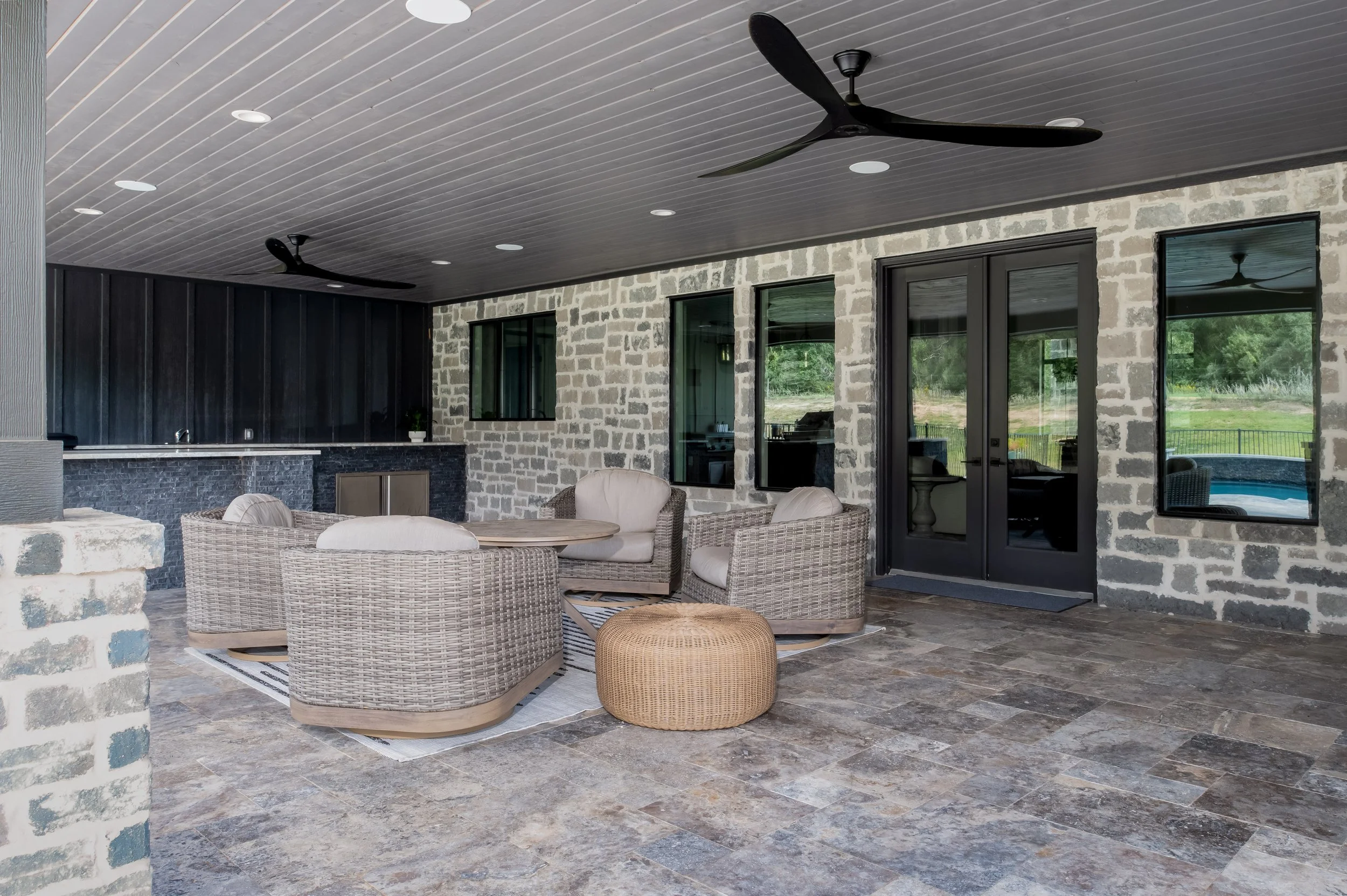 Covered outdoor patio with wicker furniture, a round table, and a pouf, with a stone wall, black door, and windows, ceiling fans, and a view of outdoor greenery and a pool.