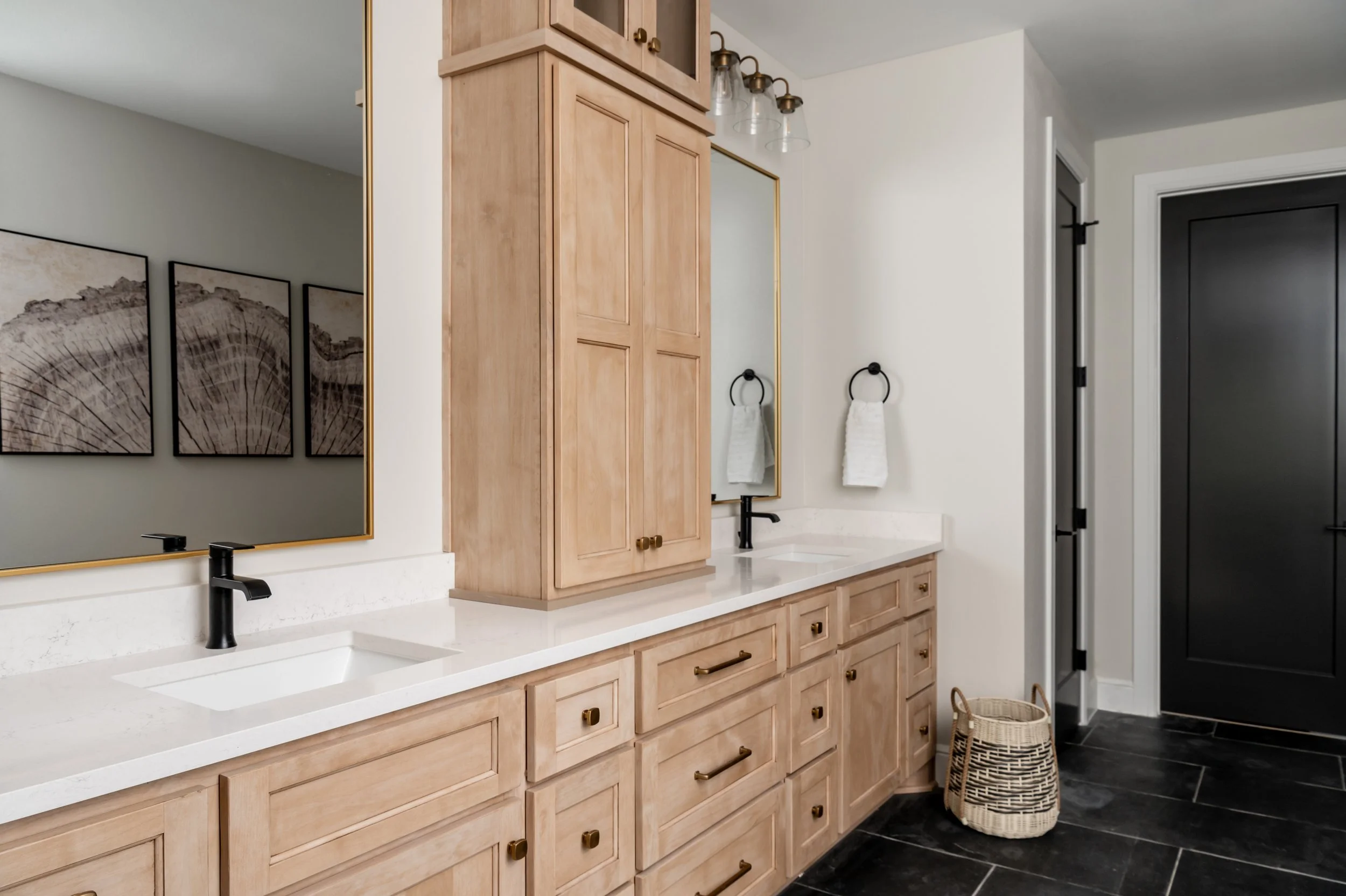 A modern bathroom vanity with light wood cabinets, a white countertop, and black faucets. There are two sinks, a large mirror, and black towel rings with white towels. A black door is visible in the background, and a woven basket is on the dark tiled