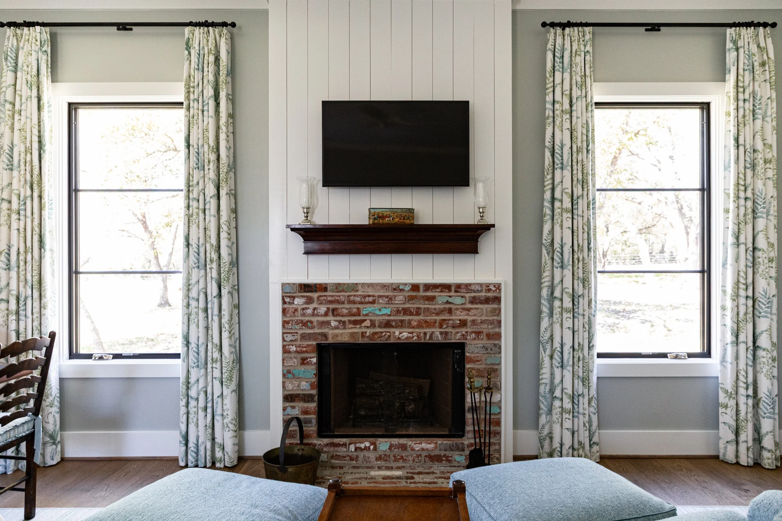 Living room with a fireplace, brick surround, and a mounted flat-screen TV above it. Two windows with floral curtains flank the fireplace, with trees visible outside. A wooden shelf above the fireplace holds decorative items and lamps. Part of a blue