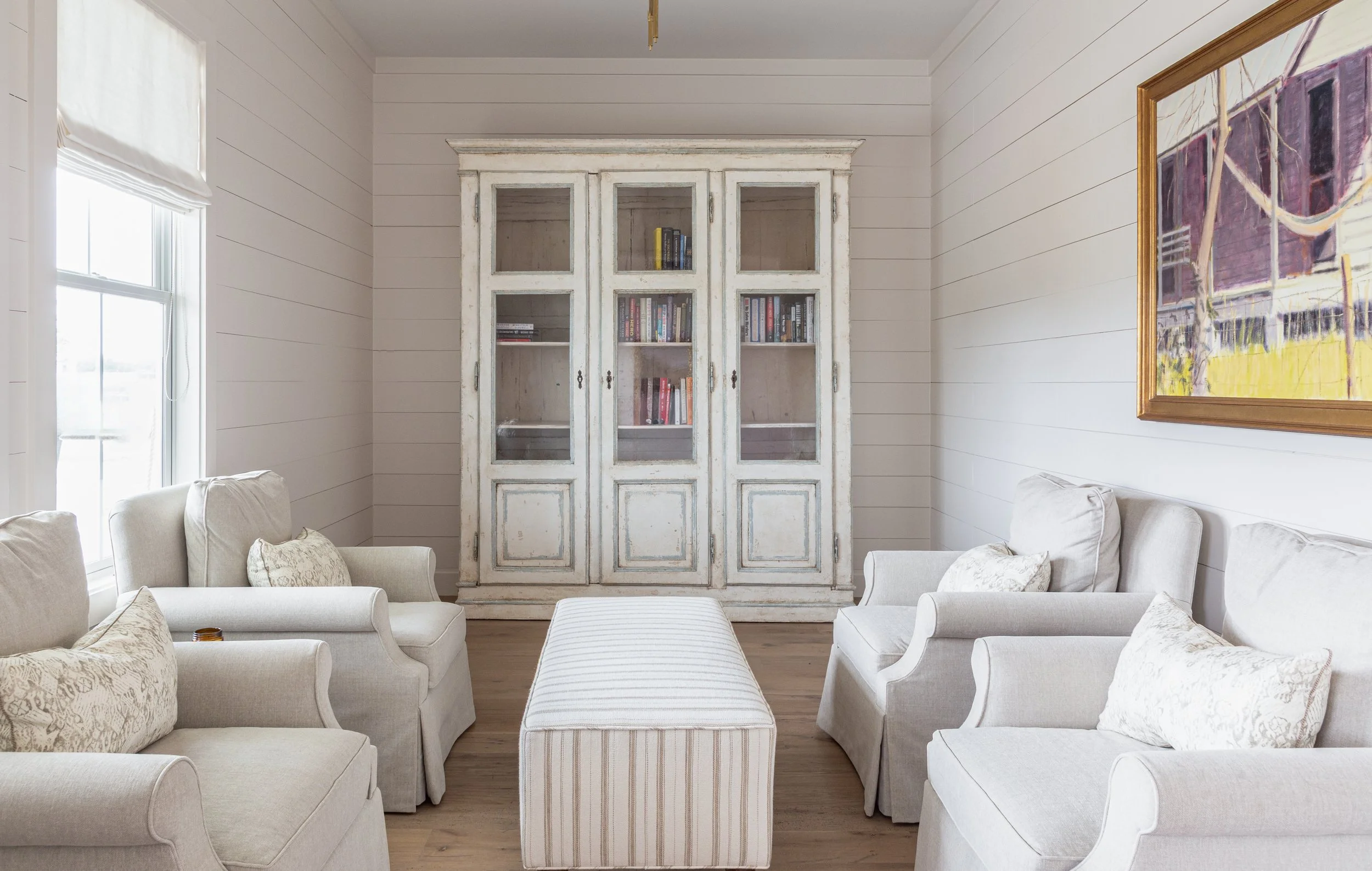 Living room with white sofas, a striped ottoman, a white distressed cabinet with glass doors, a window with a white blind, and a framed painting on the wall.