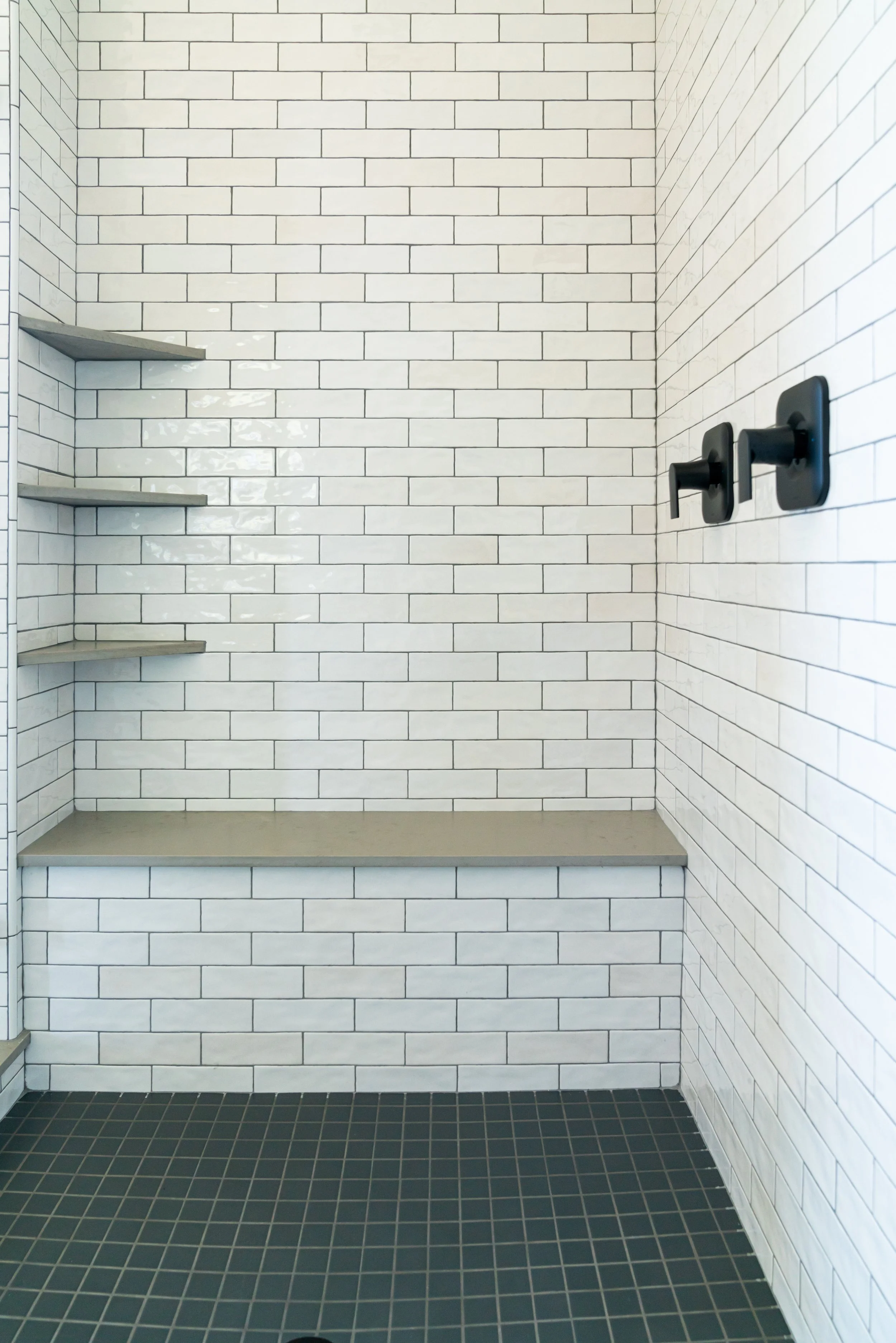 Empty shower with white subway tile walls, black shower handles, and gray shelves.
