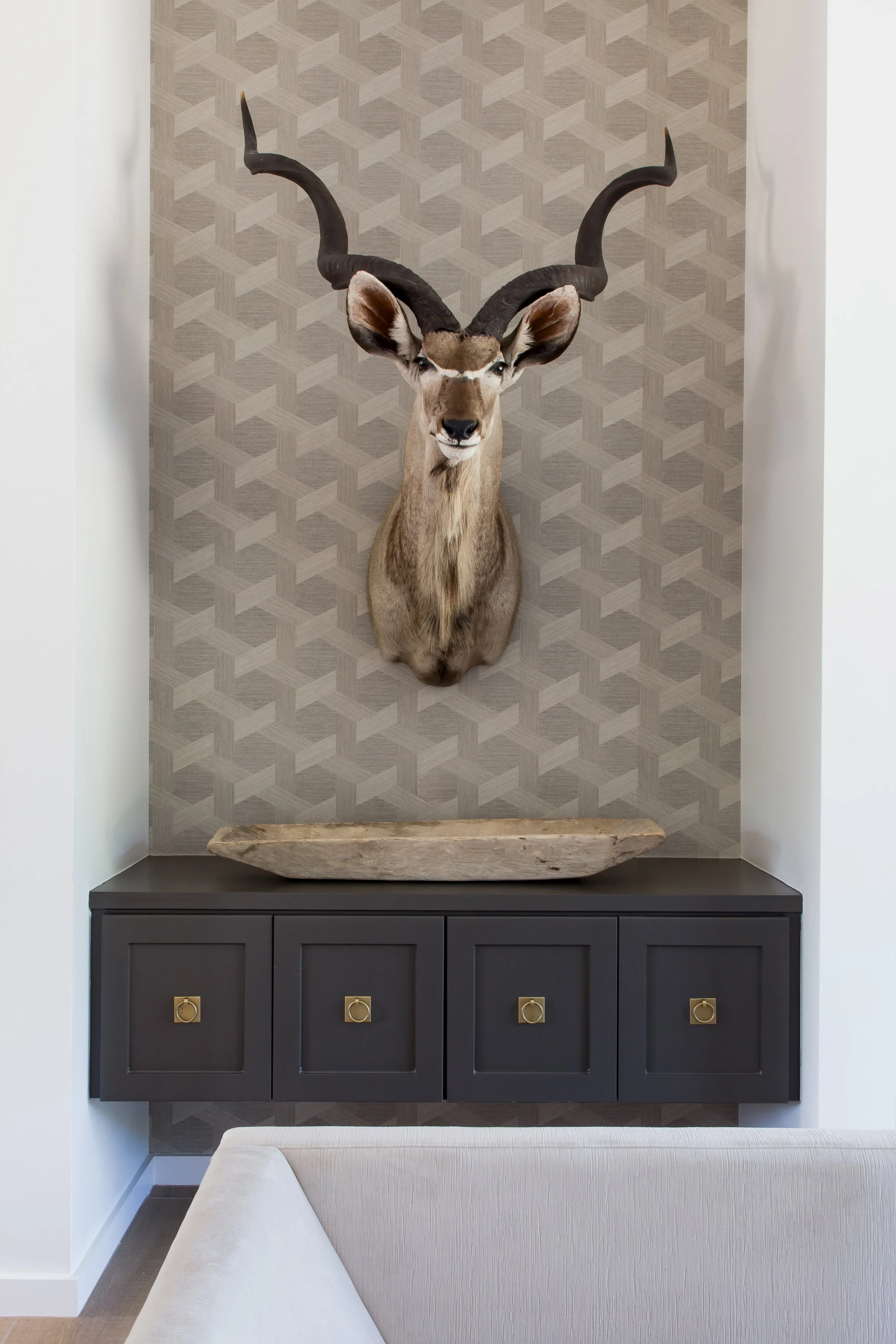 Mounted animal head, possibly a deer or an antelope, with large curved horns on a textured wallpaper background, positioned above a black cabinet with gold handles and a wooden tray.