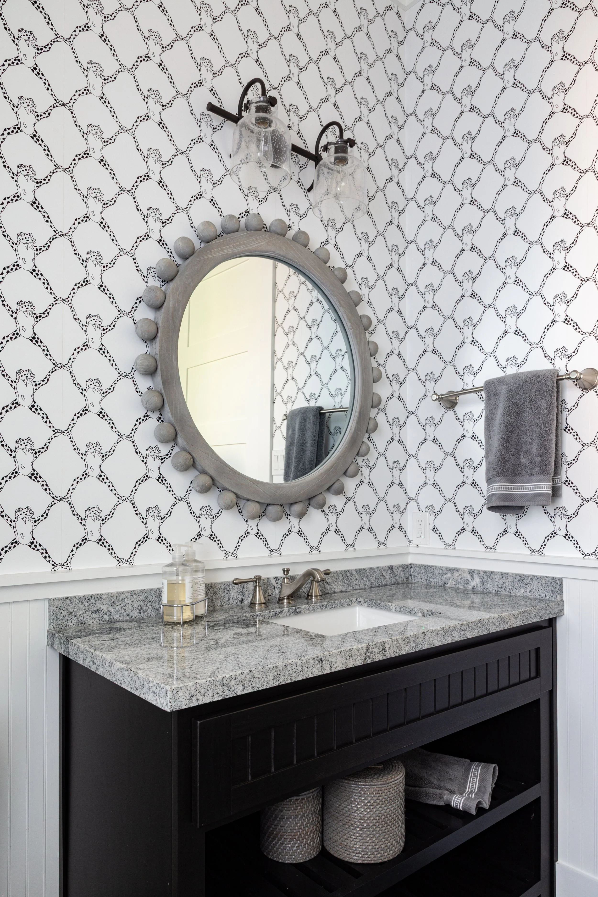Bathroom with black and white cheetah print wallpaper, round mirror with decorative frame, granite countertop with sink, gray hand towel on towel bar, and gray storage baskets underneath.