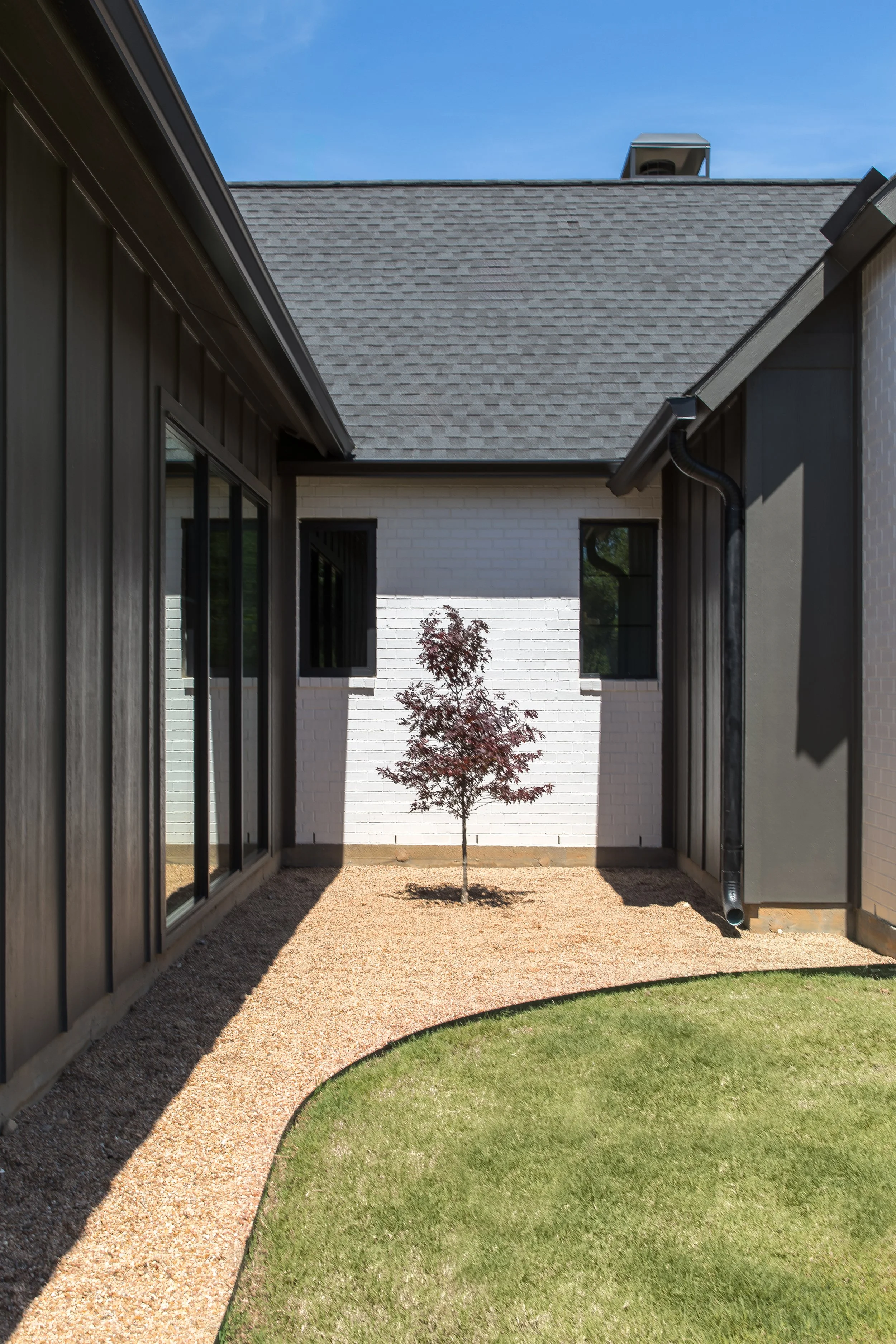 A small red tree in a backyard between two modern houses with gray and white walls, a grass patch, and gravel pathway under a blue sky.