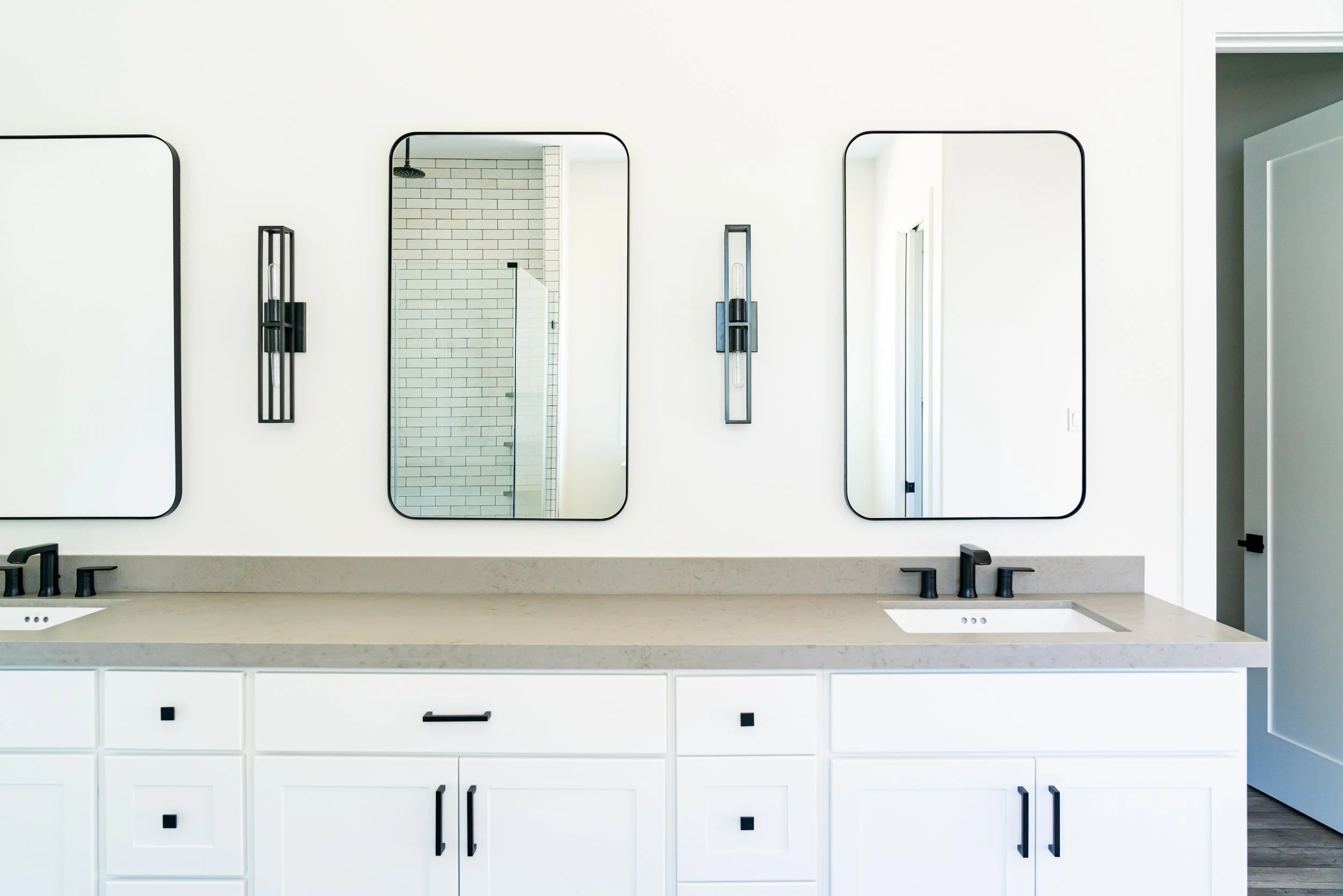 Modern bathroom vanity with three mirrors, black fixtures, and a beige countertop, reflected in the mirrors, with a view of the shower area with white subway tiles.