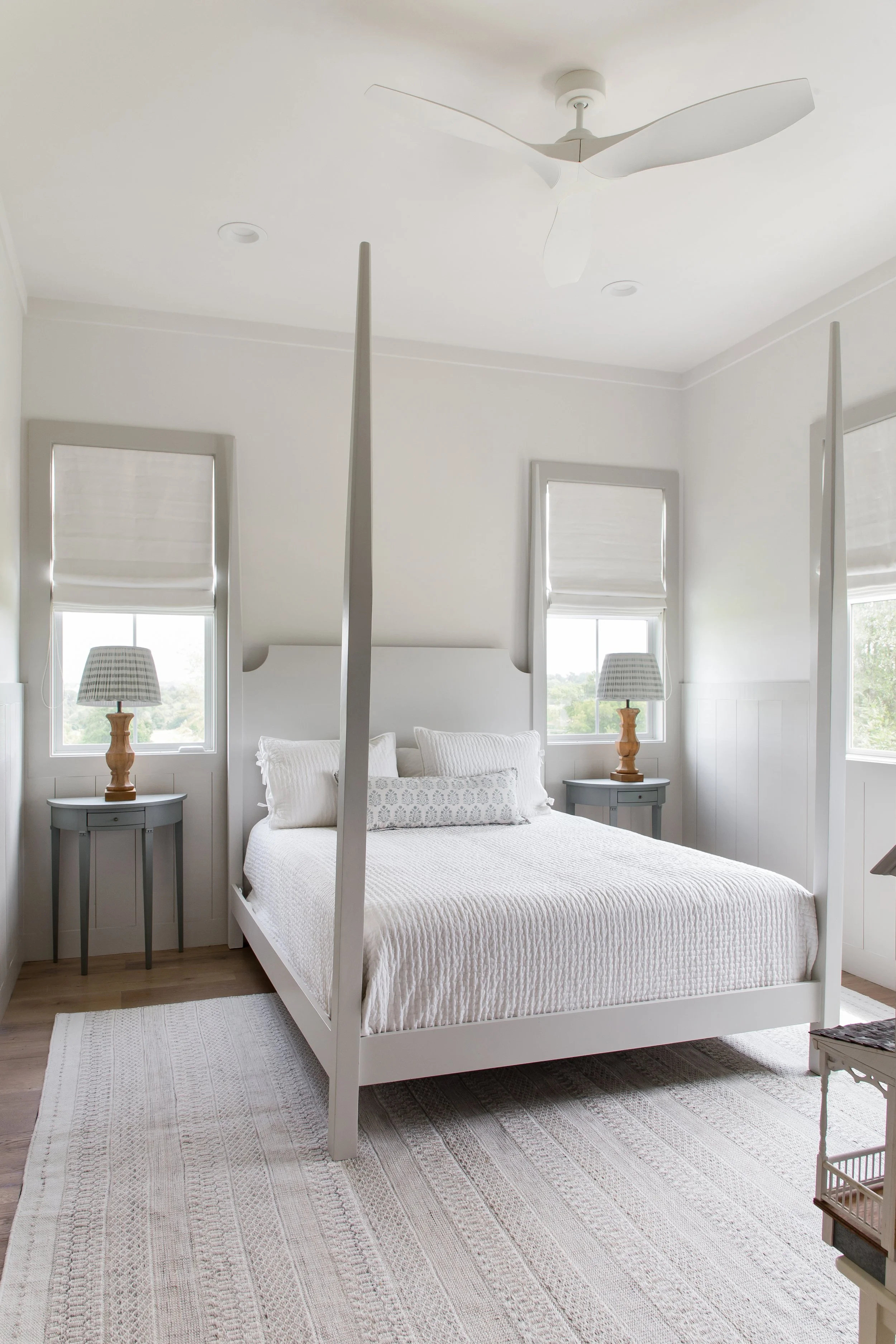 A bedroom with white walls and bedding, a white four-poster bed, two gray nightstands with lamps, two windows with white blinds, a ceiling fan, and a light-colored area rug.