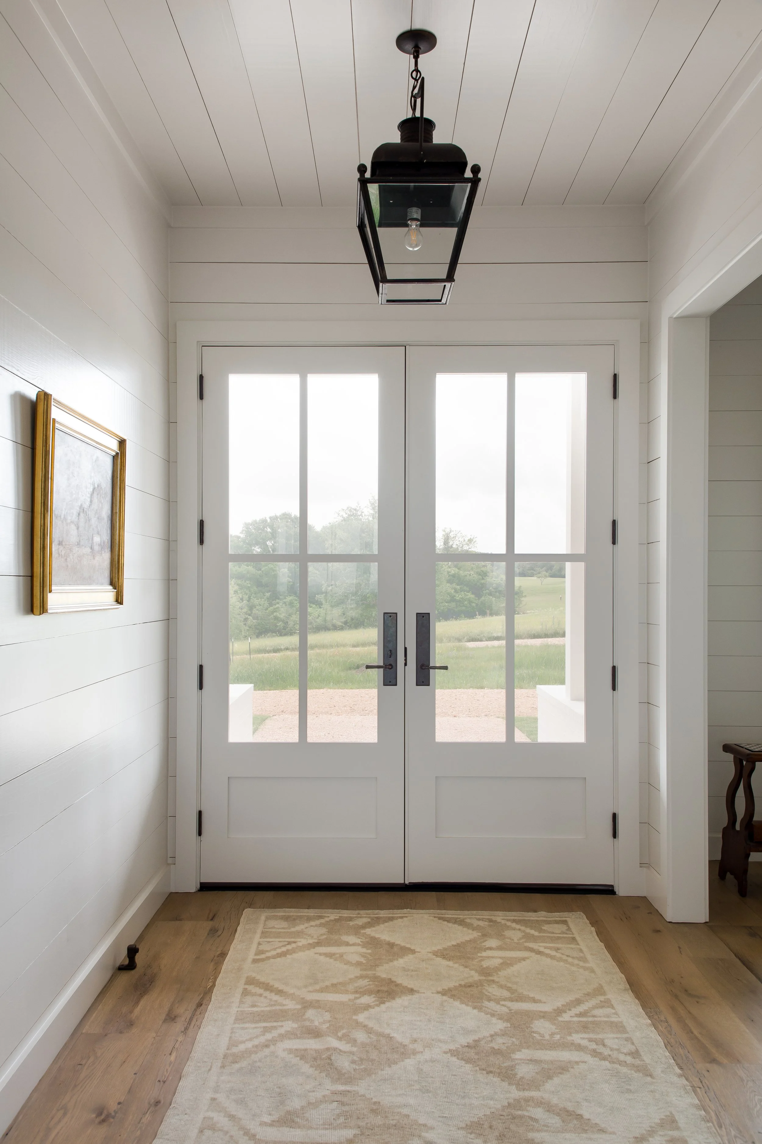 Interior view of a doorway with glass double doors leading outside, with a view of greenery and a cloudy sky, a ceiling lantern hanging above, and a patterned rug on the wooden floor.