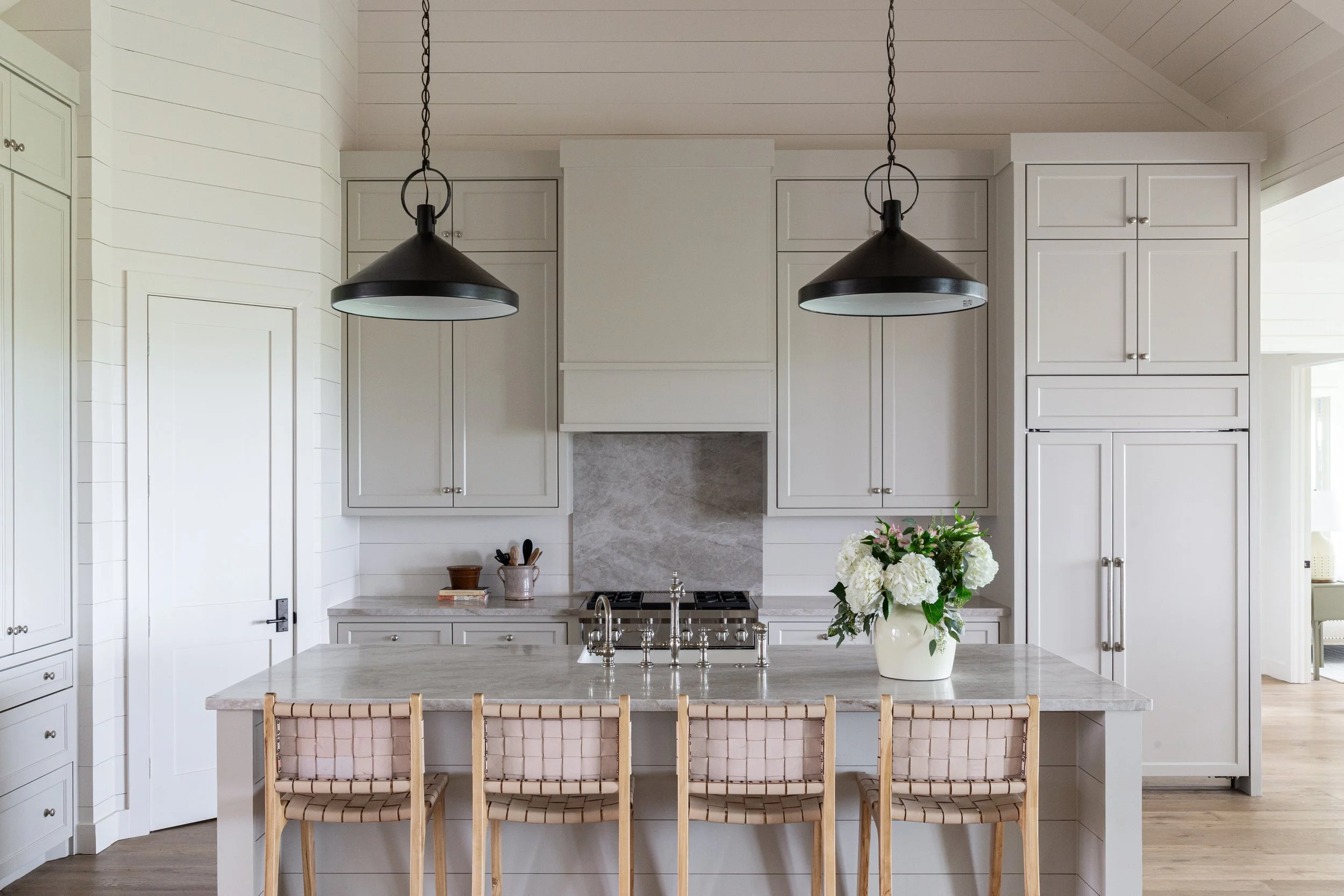 Modern white kitchen with marble island, large vase of white flowers, overhead black pendant lights, and built-in cabinets.