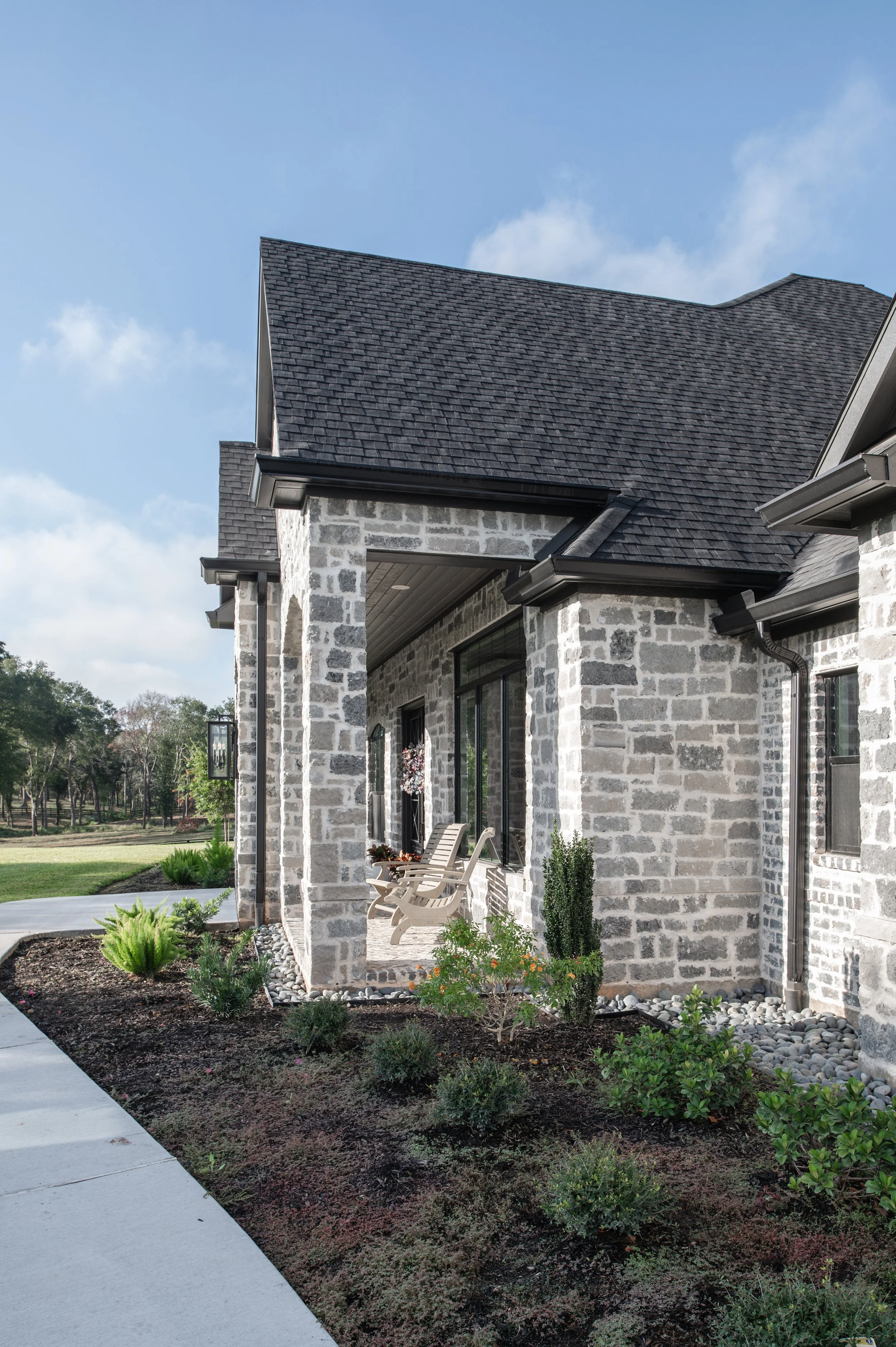 A house with stone exterior walls, black roof shingles, and a front porch with chairs surrounded by landscaped bushes and plants. A sidewalk is visible in the foreground.