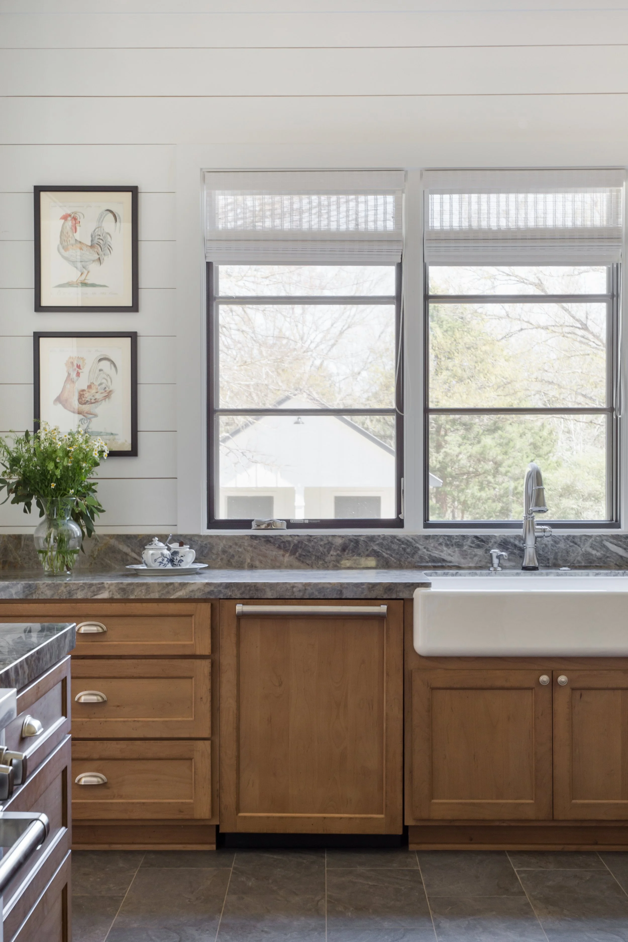 A kitchen with wooden cabinets, a marble countertop, a farmhouse sink under large window, and framed rooster pictures on the white shiplap wall.