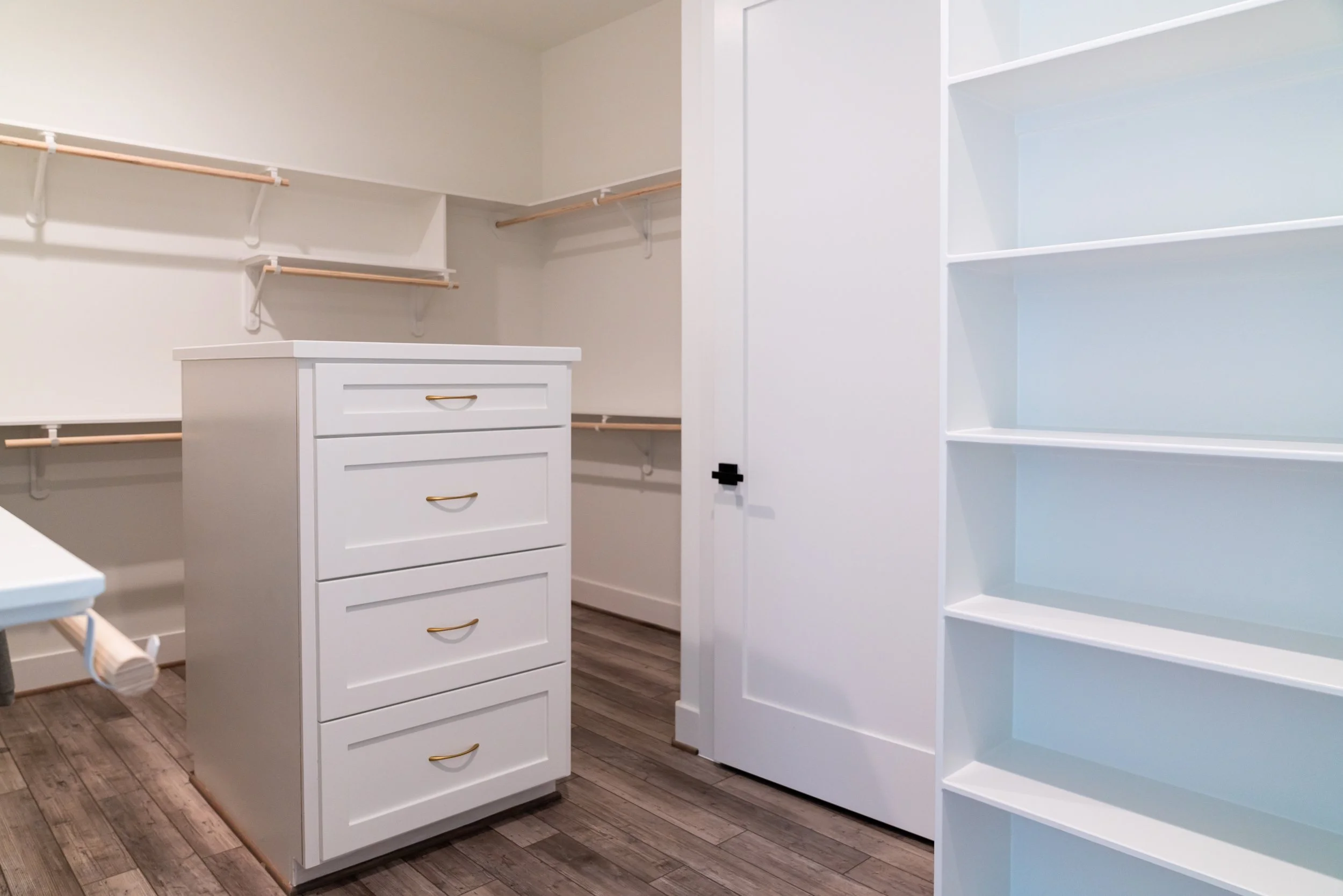 Empty walk-in closet with white shelves, drawers, and hanging rods, with wood flooring.