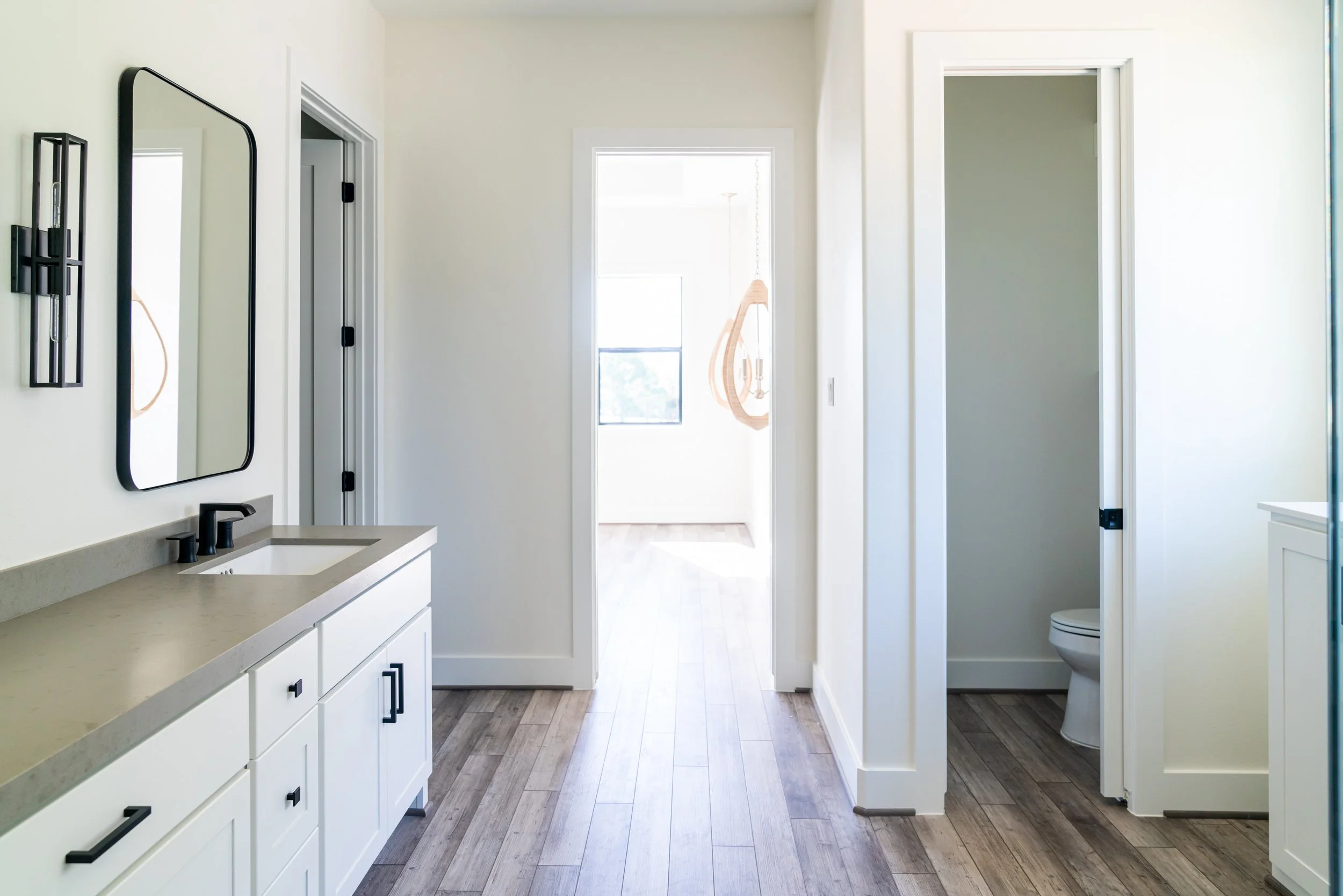 Bathroom with white cabinetry, a black-framed mirror, and two doors leading to a toilet and a bright room with a window and hanging art.