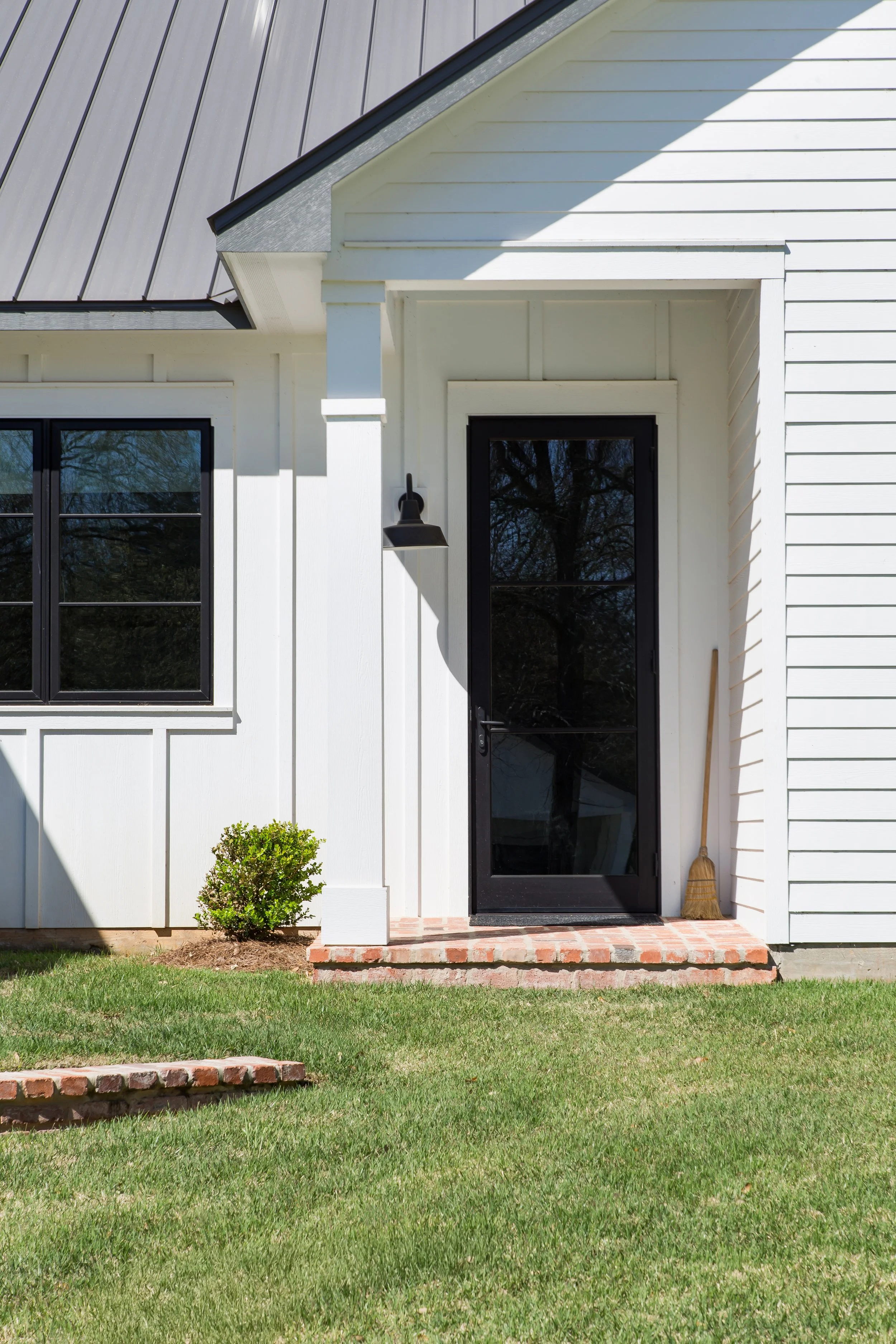 Exterior of a white house with a black glass door and window, small porch steps, a small bush, a wall-mounted light, and a broom leaning against the house