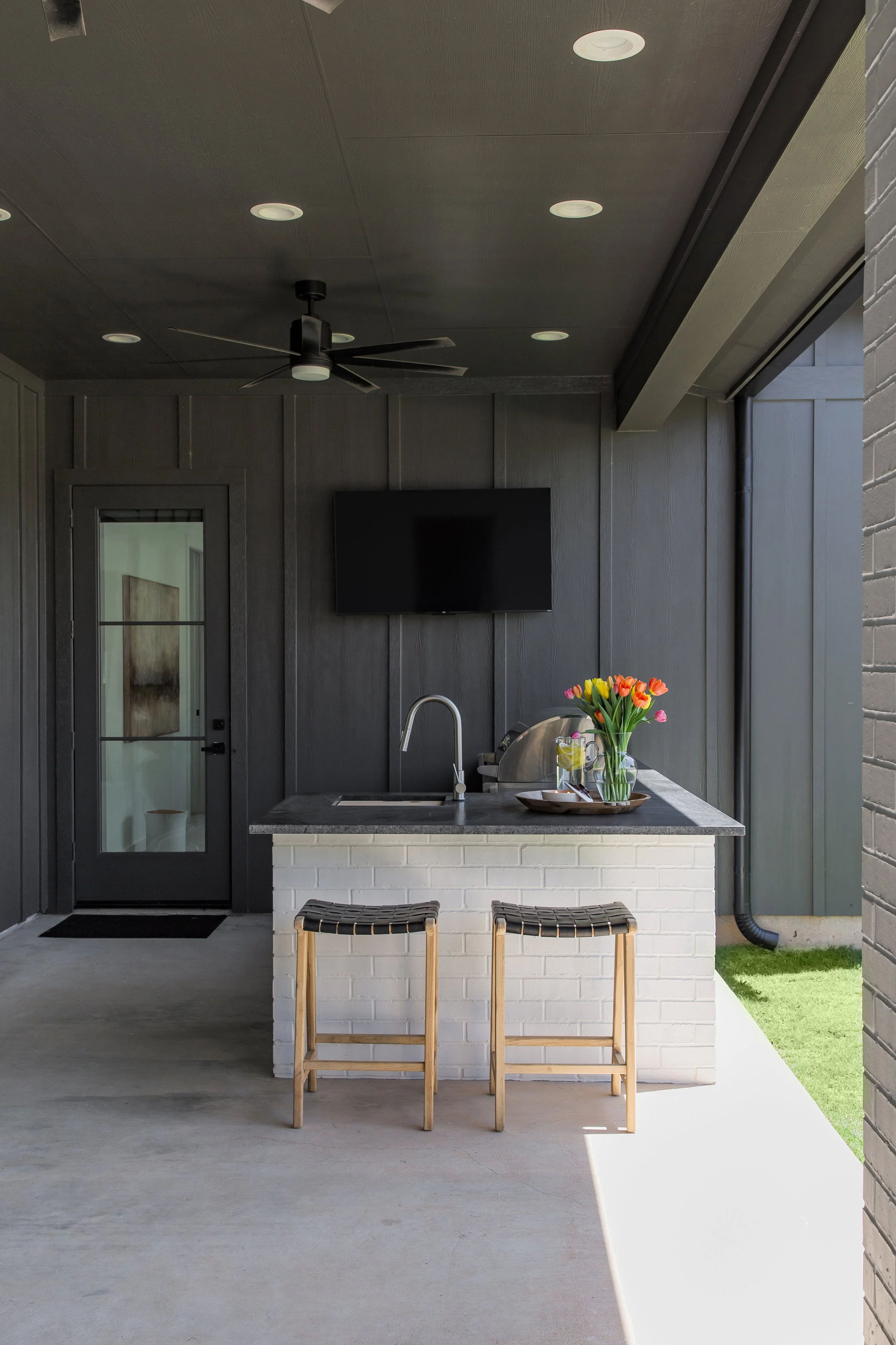 Modern outdoor kitchen with a black ceiling, dark gray walls, and a white brick island topped with a flower vase and tray, featuring two wooden stools in front of the island, opened to a backyard with grass.