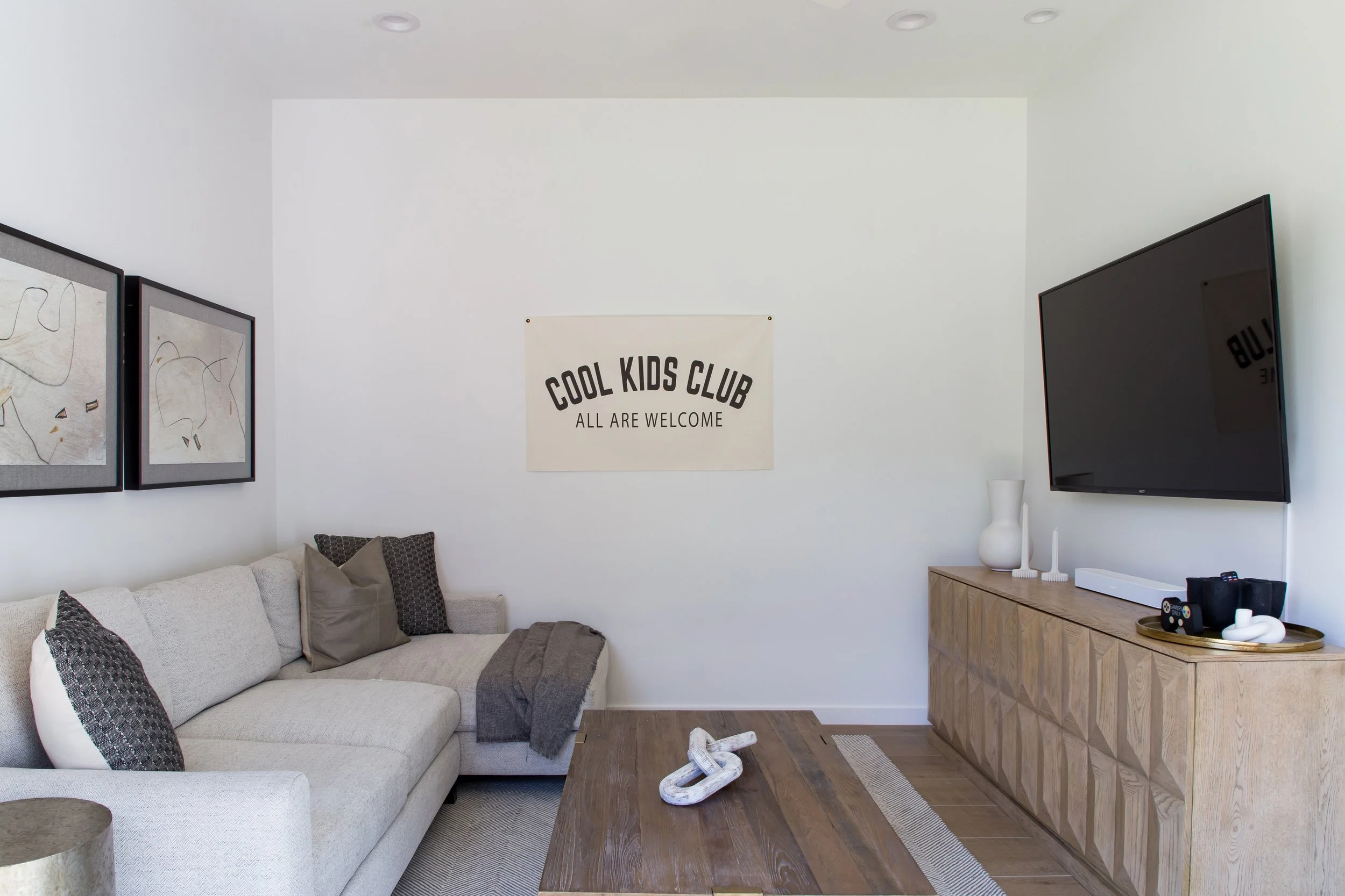Living room with a beige couch, a wooden coffee table, a wooden media console with decorative vases, a wall-mounted television, abstract artwork, and a banner that reads 'Cool Kids Club, All Are Welcome', in a modern interior.