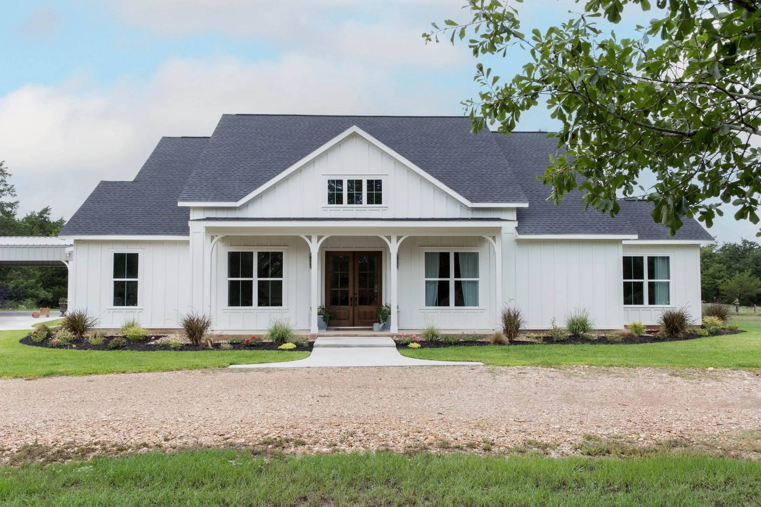 Front view of a modern white farmhouse with dark shingles, a small covered porch, and garden beds with plants and flowers.