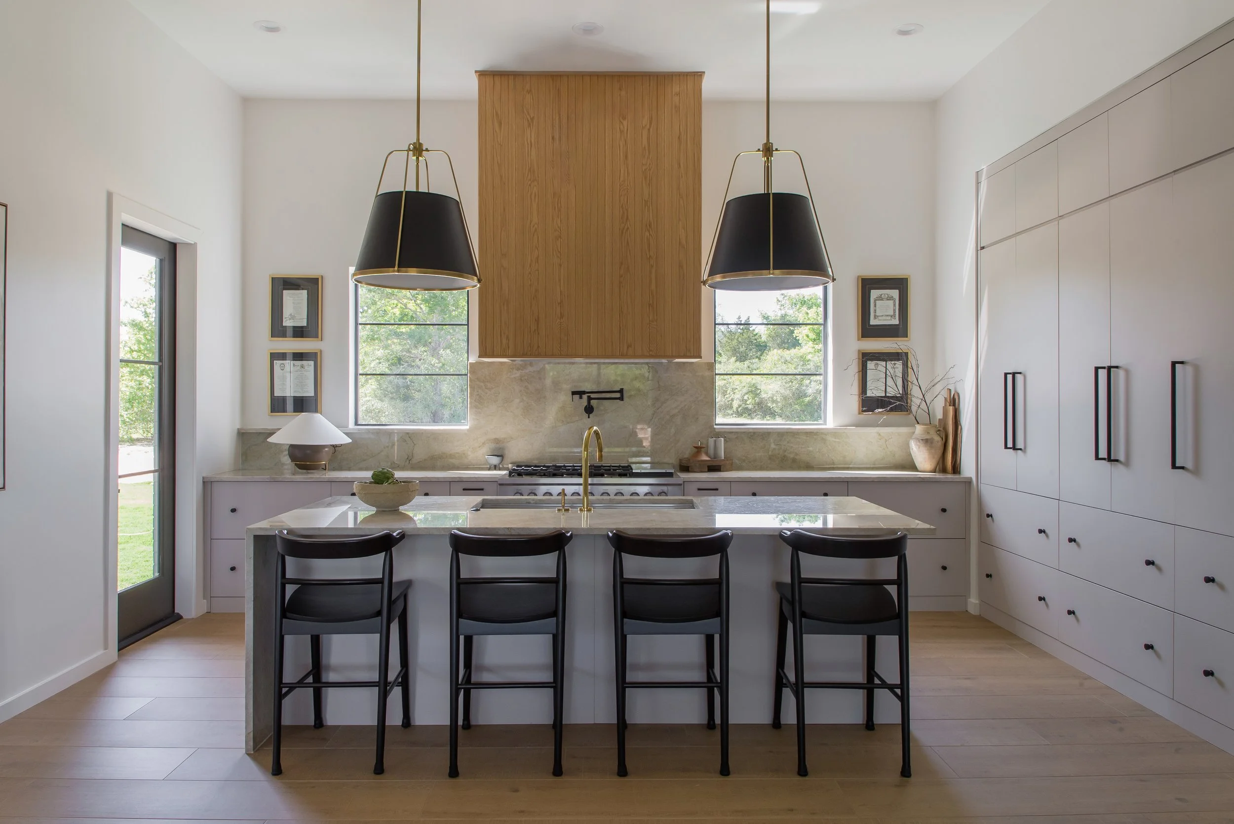 Modern kitchen with white cabinets, a central island with black bar stools, black hanging pendant lights, large windows, and minimal decor.