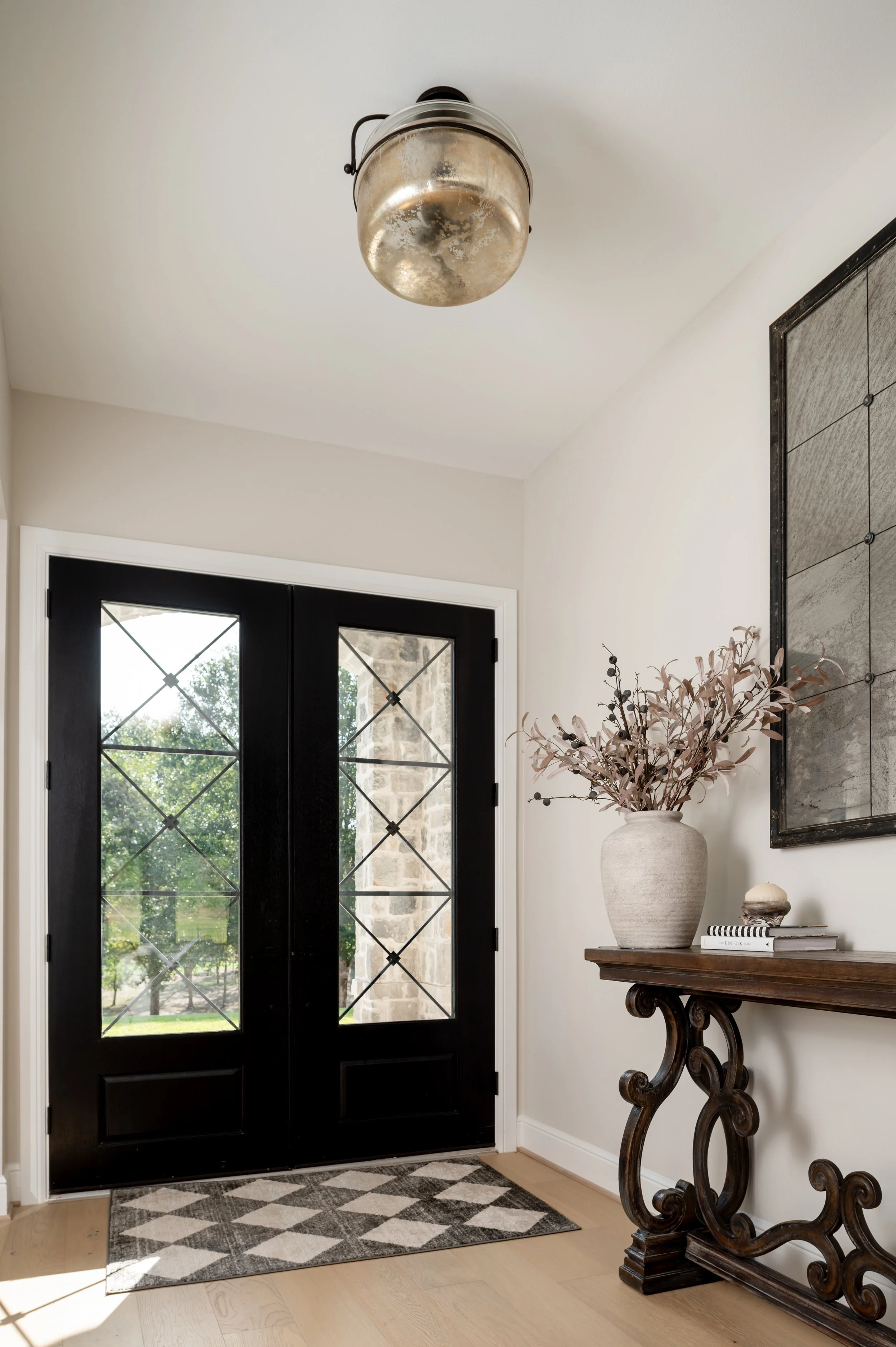 Entryway with black double front doors with decorative glass panels, a striped black and white rug, a wooden console table with a large vase of dried branches, and a framed mirror on a white wall.