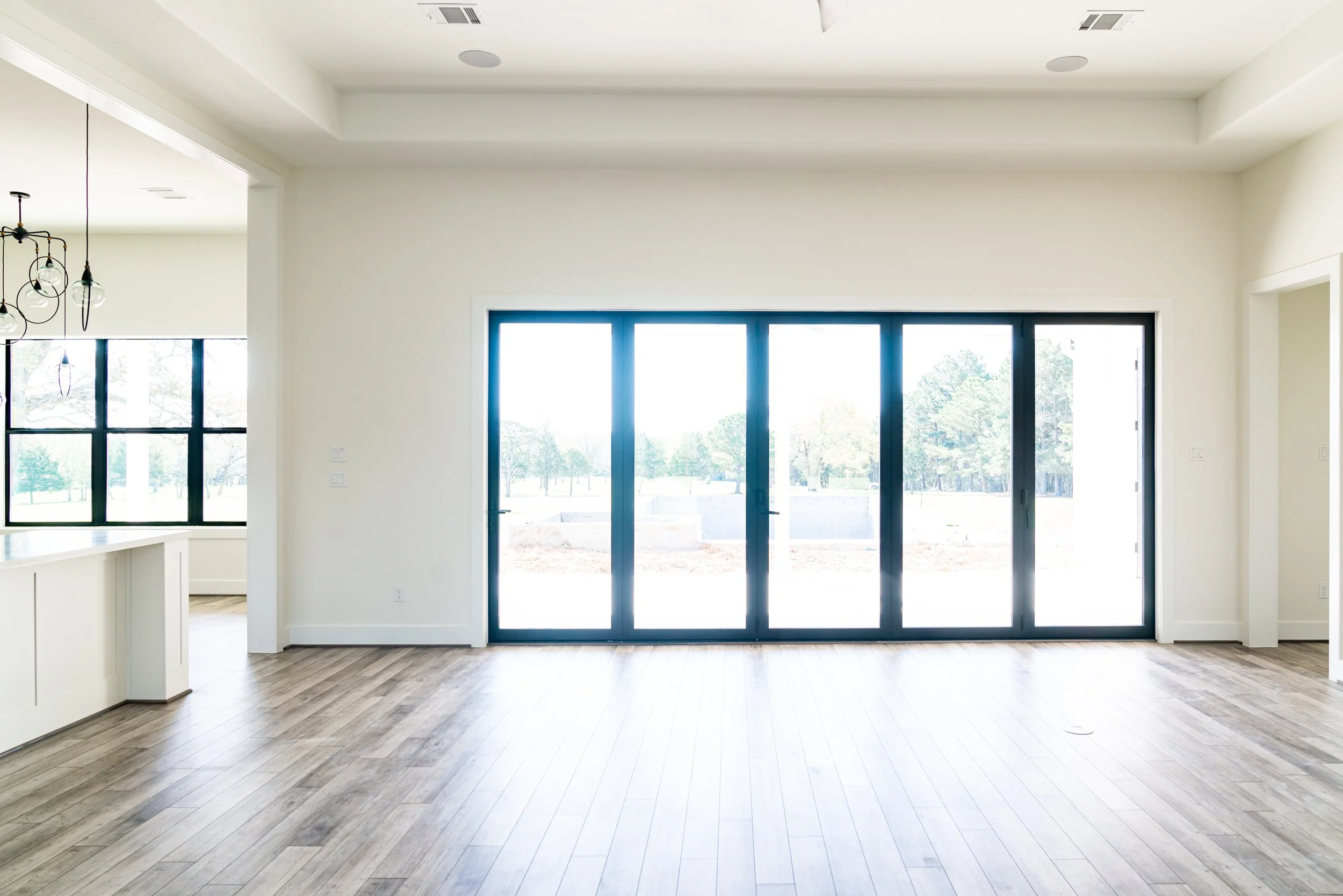 Empty living room area with large sliding glass doors leading outside, light-colored hardwood flooring, white walls, and a visible part of a kitchen with black-framed windows on the left.