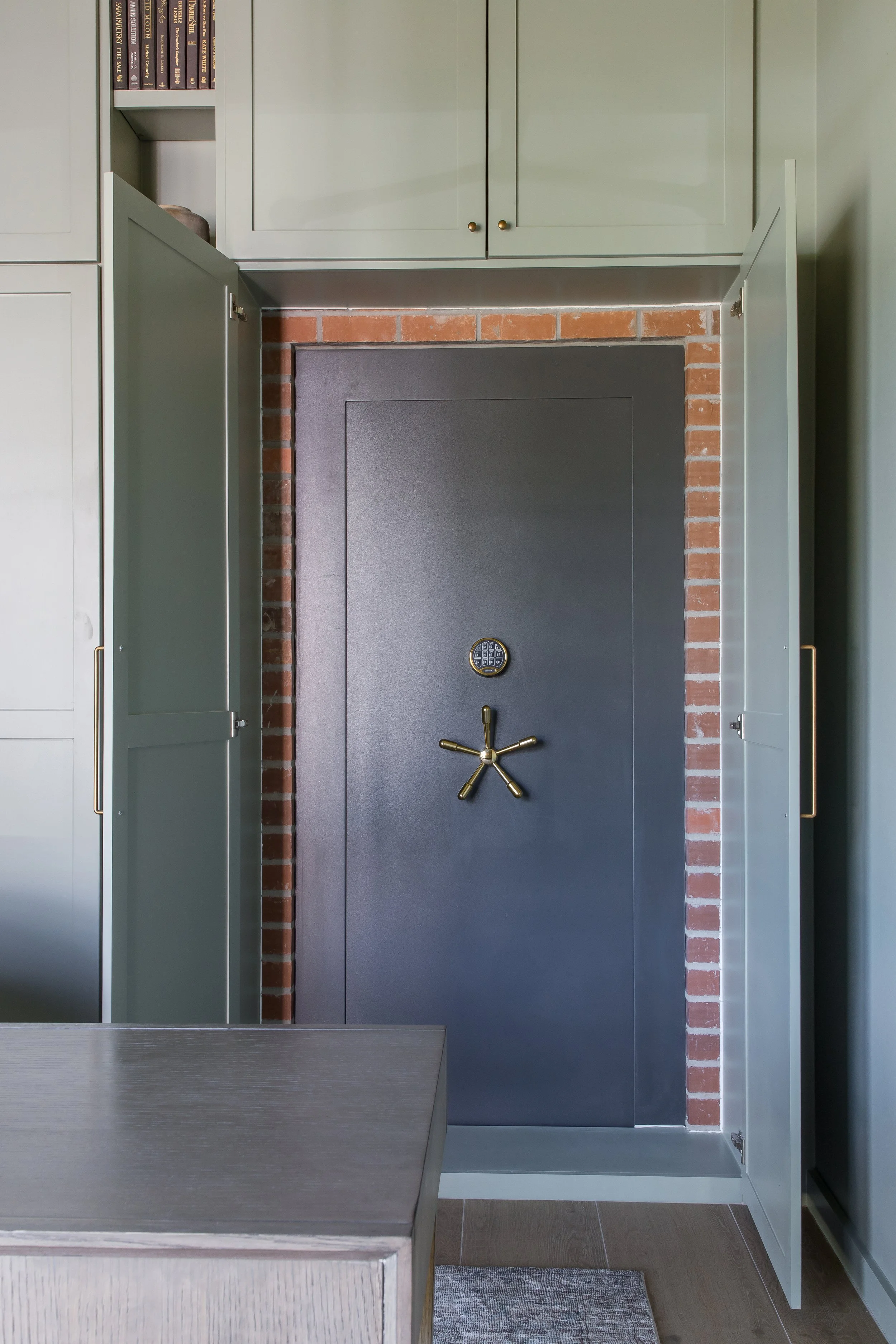 A black safe with a combination lock and handle, set in a room with green cabinets and a brick wall background.