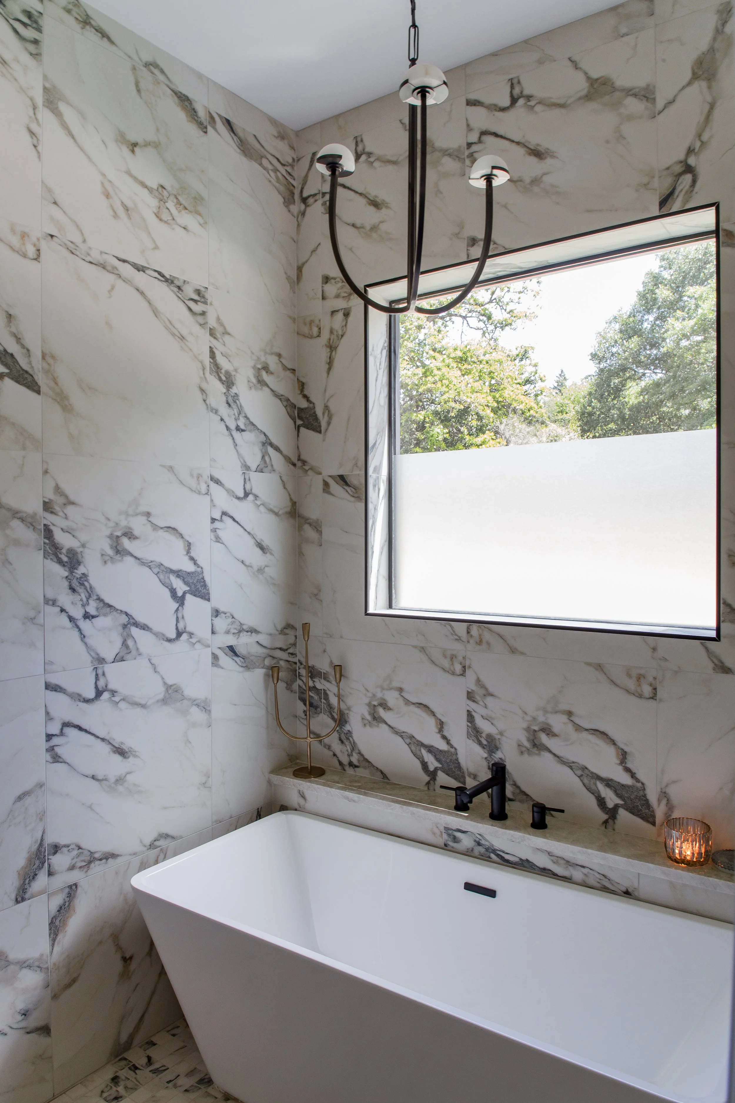 A modern bathroom with marble walls, a freestanding bathtub, a chandelier, and a large window showing trees outside.