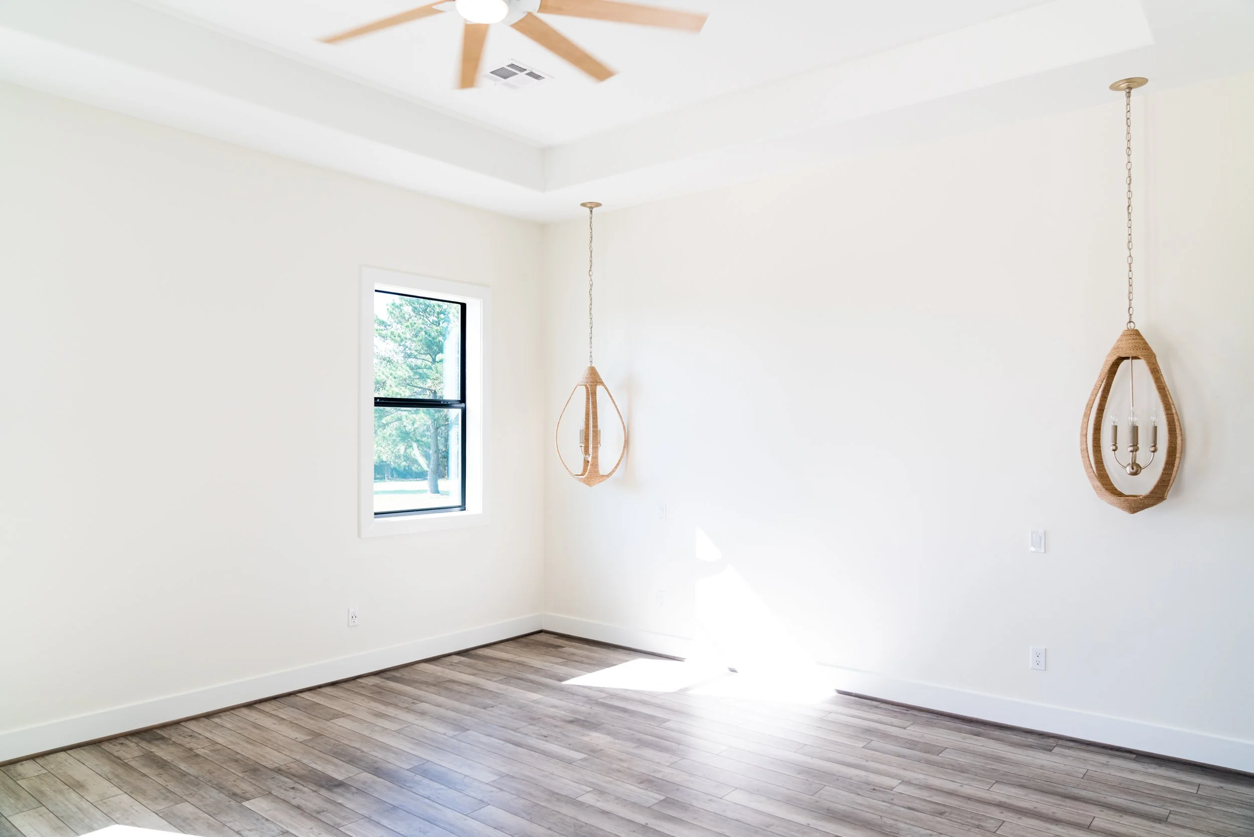 Empty room with white walls, hardwood flooring, two hanging light fixtures, a ceiling fan, and a window with trees outside.